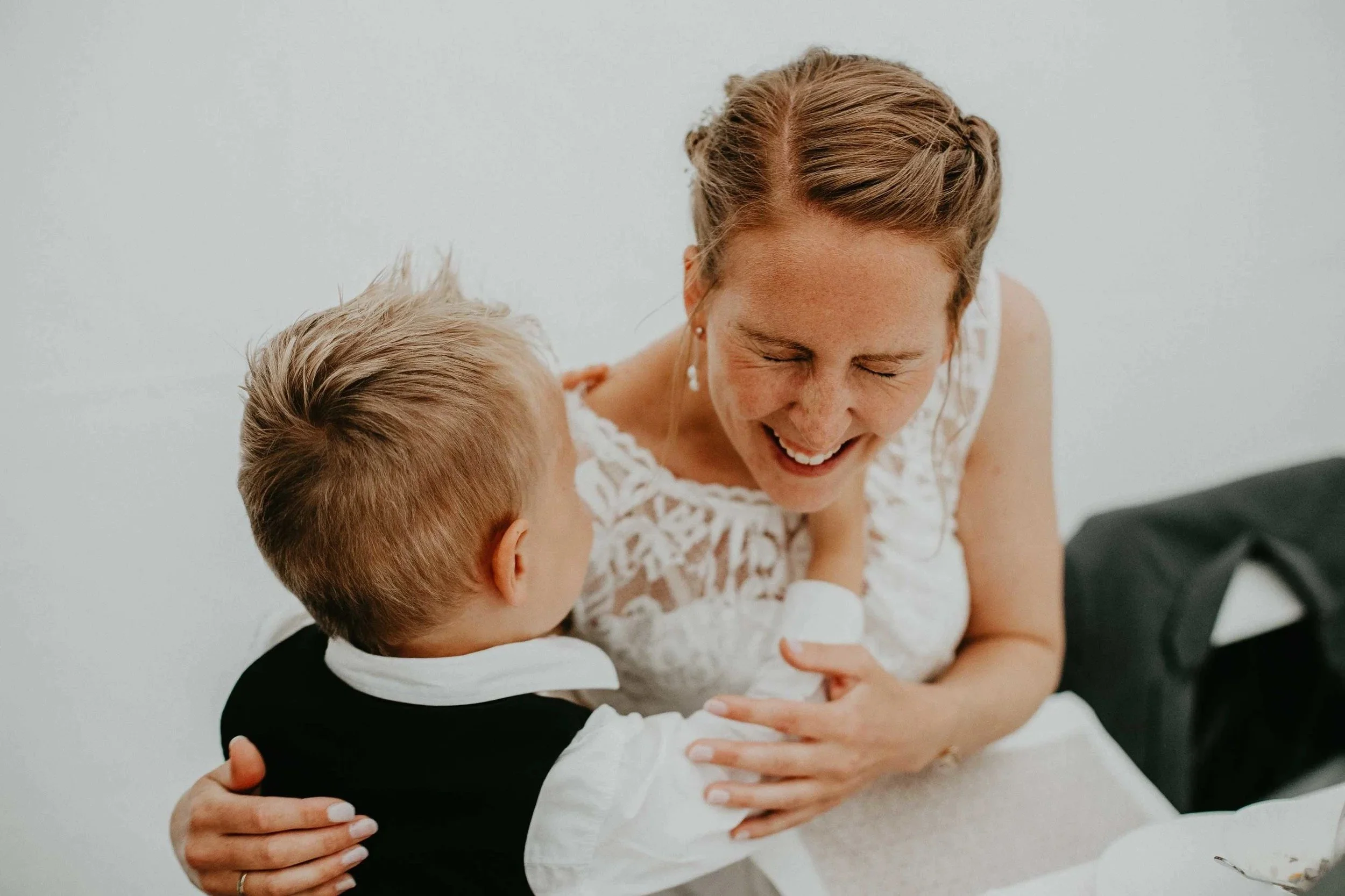 En kvinne i hvit blondekjole smiler og klemmer en ung gutt i dress.