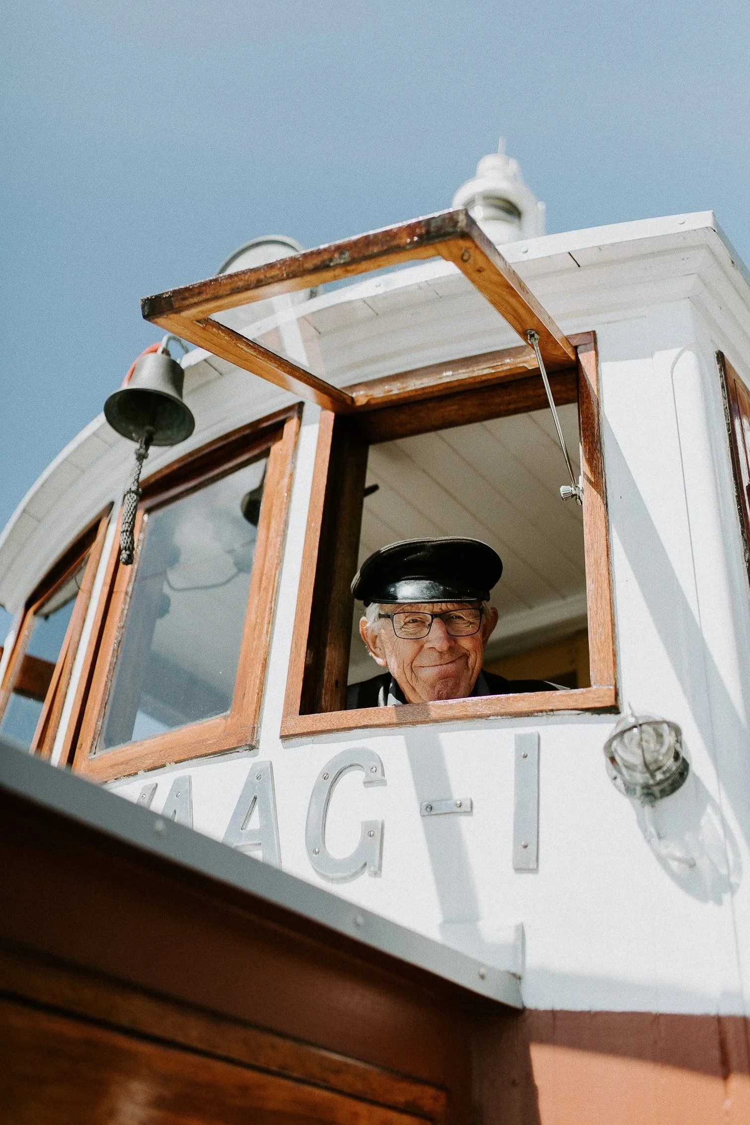 En eldre mann med kapteinslue ser ut av et båtvindu, med bjeller utenfor og blå himmel i bakgrunnen.