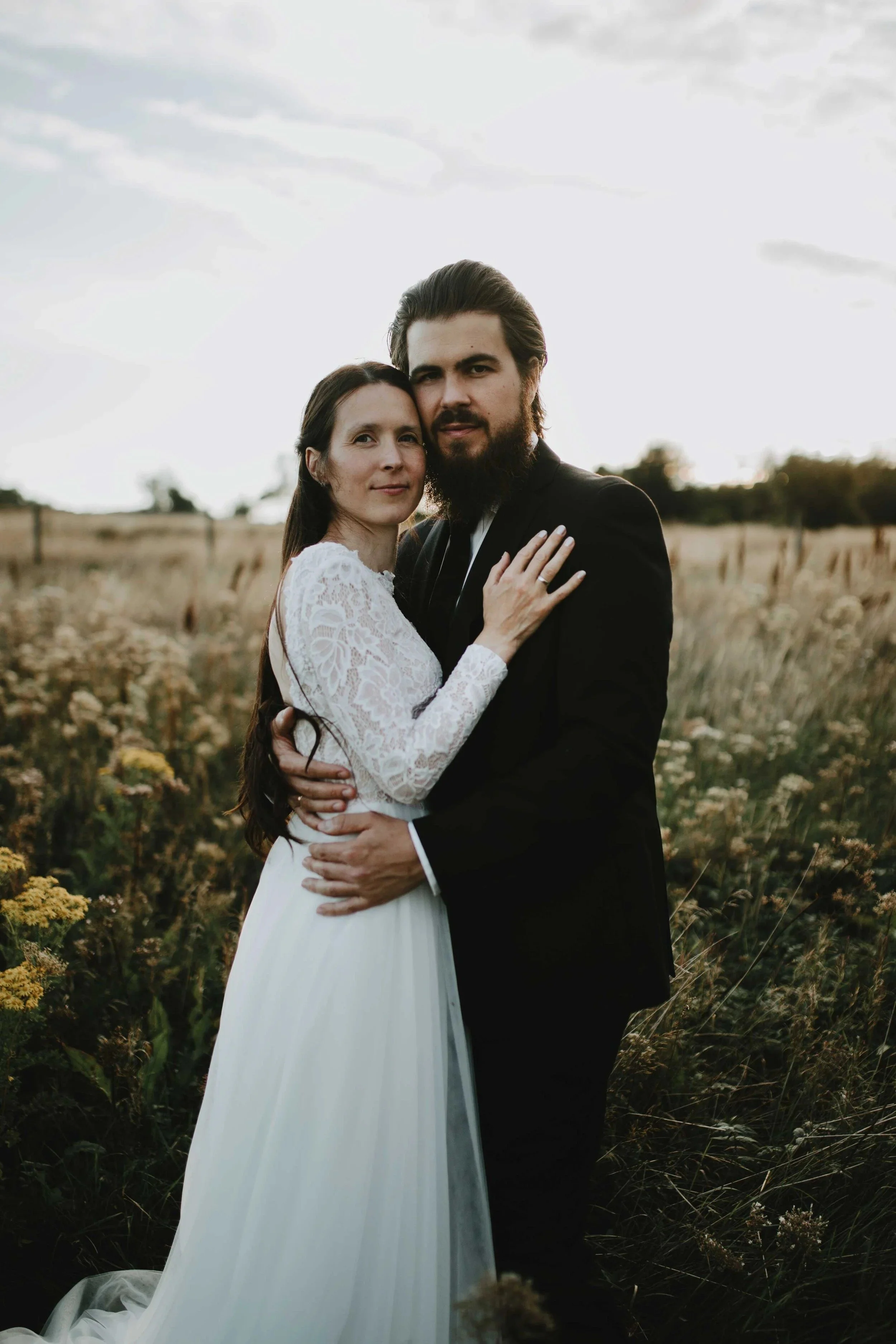 Et brudepar står tett sammen i et blomsterfelt i solnedgang, iført bryllupsantrekk.