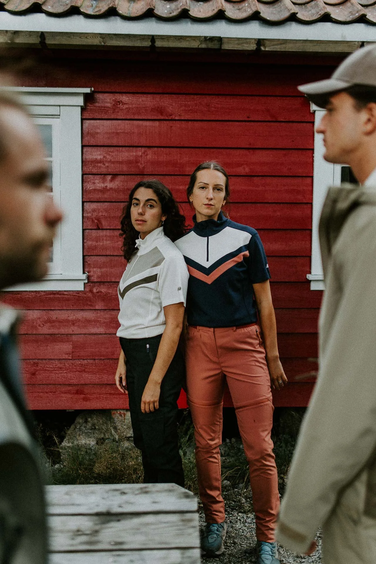 portrett av to kvinner som ser i kameraet foran rødt bygg to menn blury i forgrunnen ser inn i bildet Reklamefotografering av modeller til kampanje for Skogstad bilder tatt i preikestolenområdet bilder av turklær