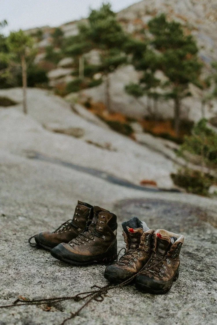 Et par godt brukte fjellsko plassert på en stein, med et uskarpt naturlandskap av trær og steinete terreng i bakgrunnen.