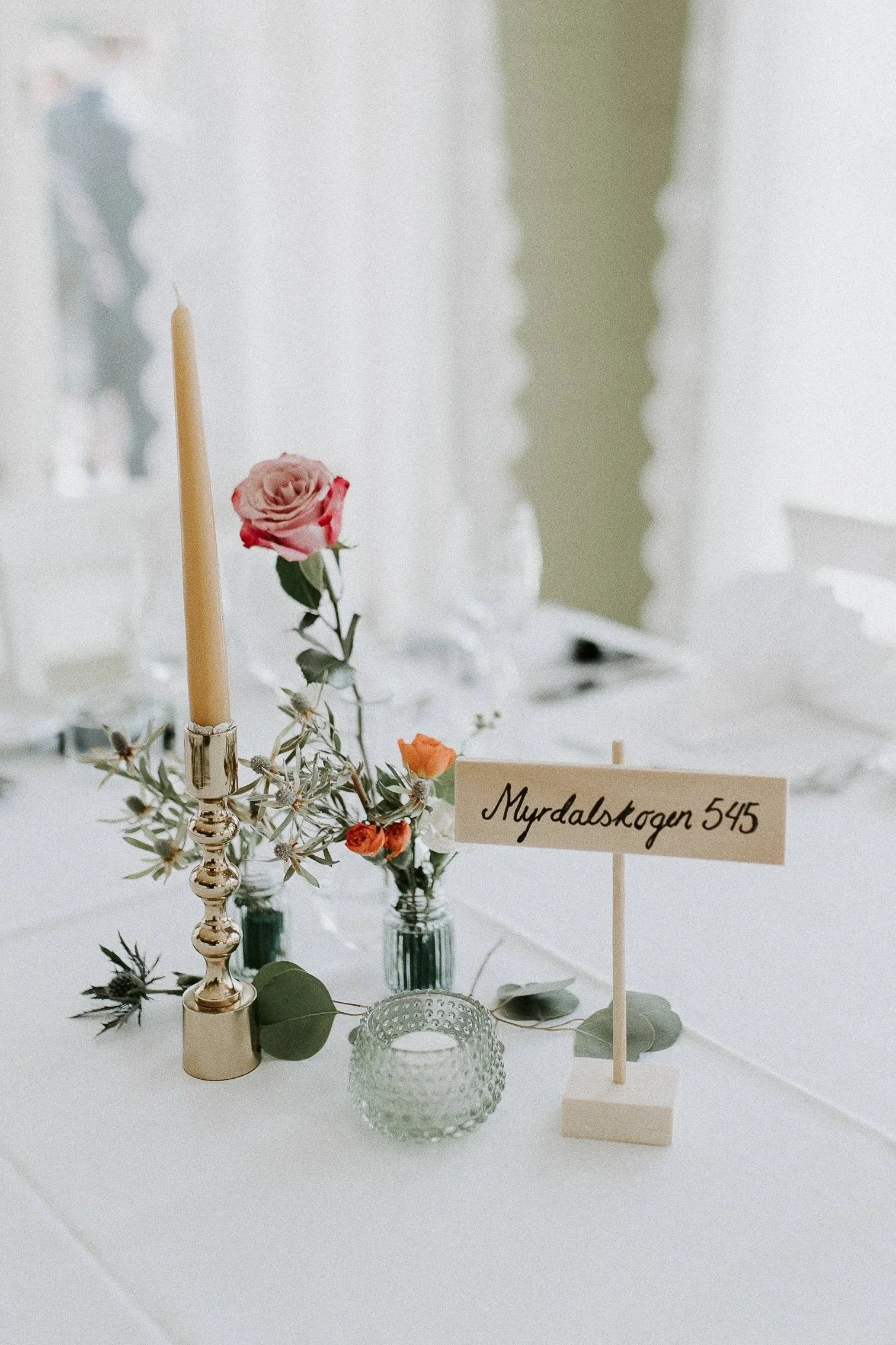 Dekorativ borddekorasjon med rosa og oransje blomster, et høyt stearinlys, grønn lysestake og håndskrevet bordkort med teksten Myrdalskogen 545.