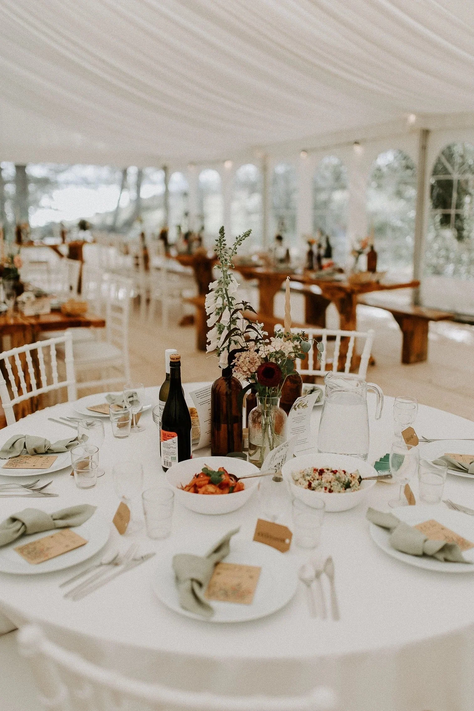 Elegant borddekking i bryllupstelt med hvite tallerkener, vinflasker, glass, servietter og bordkort. Blomster arrangert i flasker som midtpunkt.