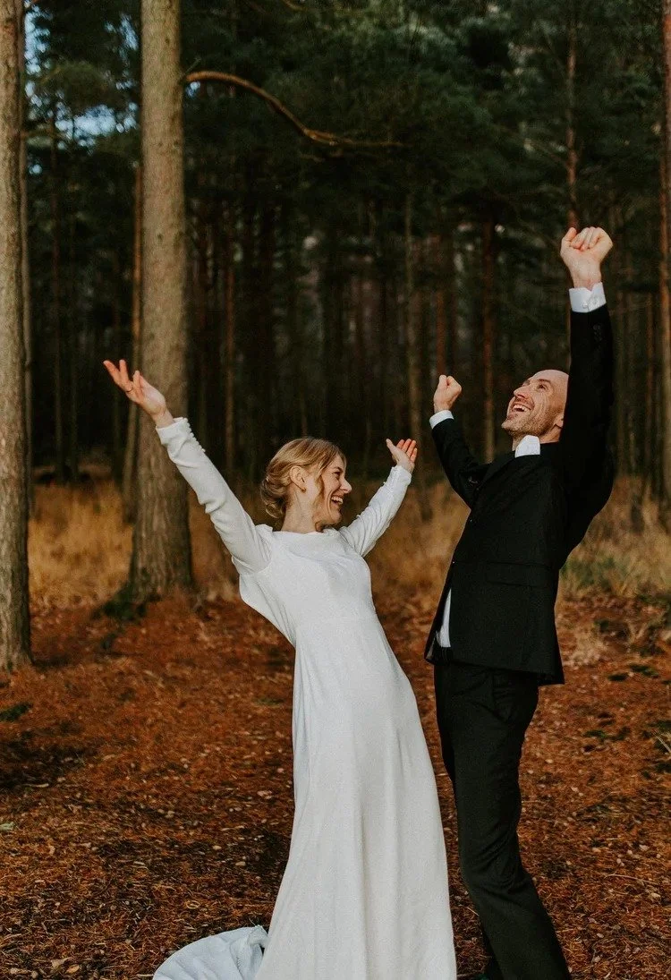 Et brudepar i bryllupsantrekk feirer med armene hevet i et skogsområde med høye trær og høstløv på bakken.