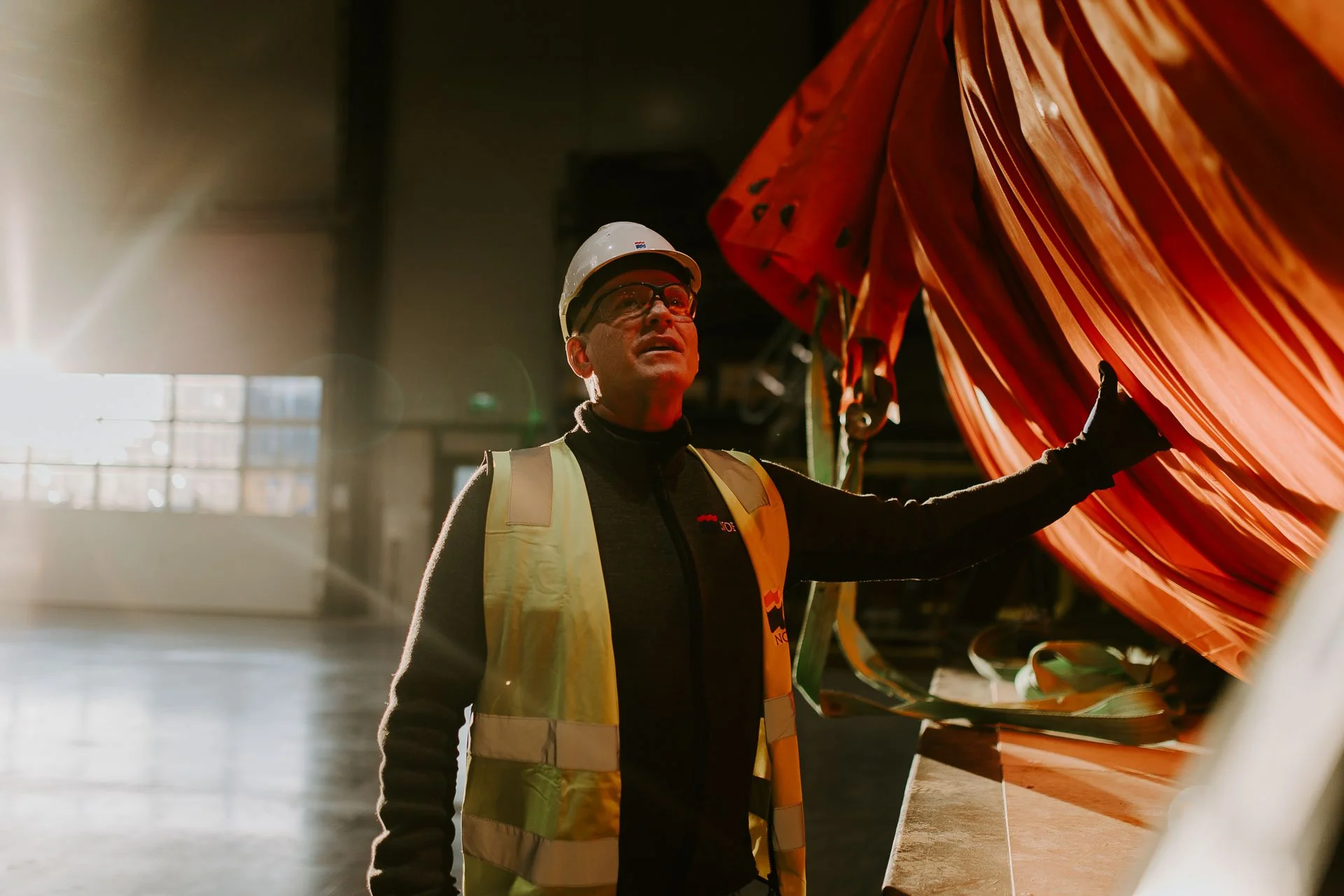 Mann som står inne i verksted med vernebriller på hvit hjelm har på gul refleksvest med nofologo holder på en oransje trommel lavt lys kommer inn vinduet bakfra reklamefotografering for nofo norsk oljevernberedskap tatt i naturlig lys