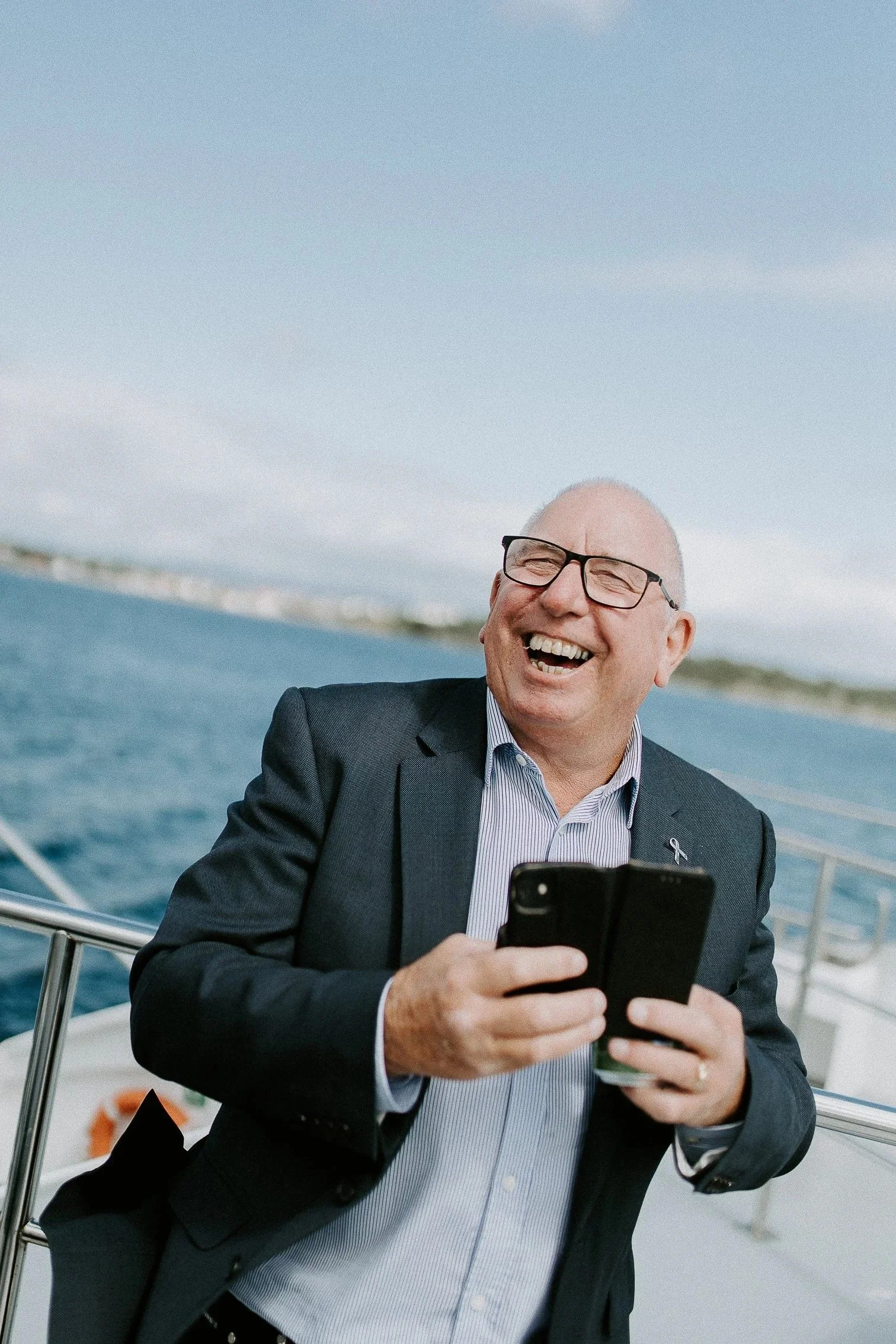 En mann i dress ler og holder en mobiltelefon på en båt, med vann og land i bakgrunnen.