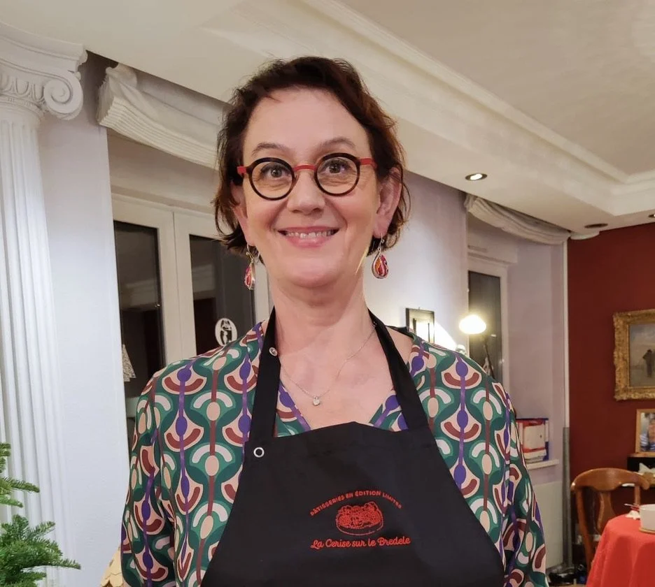 Une femme souriante portant des lunettes rondes, un tablier noir, des boucles d'oreilles pendantes, et une robe à motif coloré, dans une salle décorée.