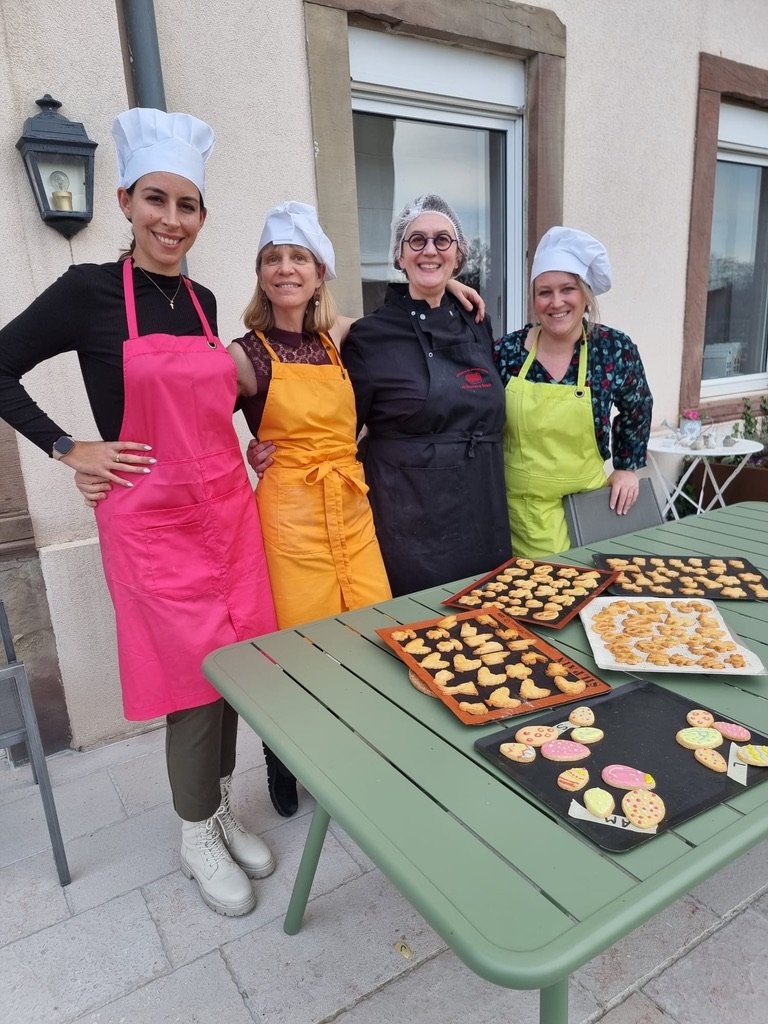groupe de quatre personnes habillé de tablier coloré, sur la table devant elles se trouve des bredele fraichement sorti du four.