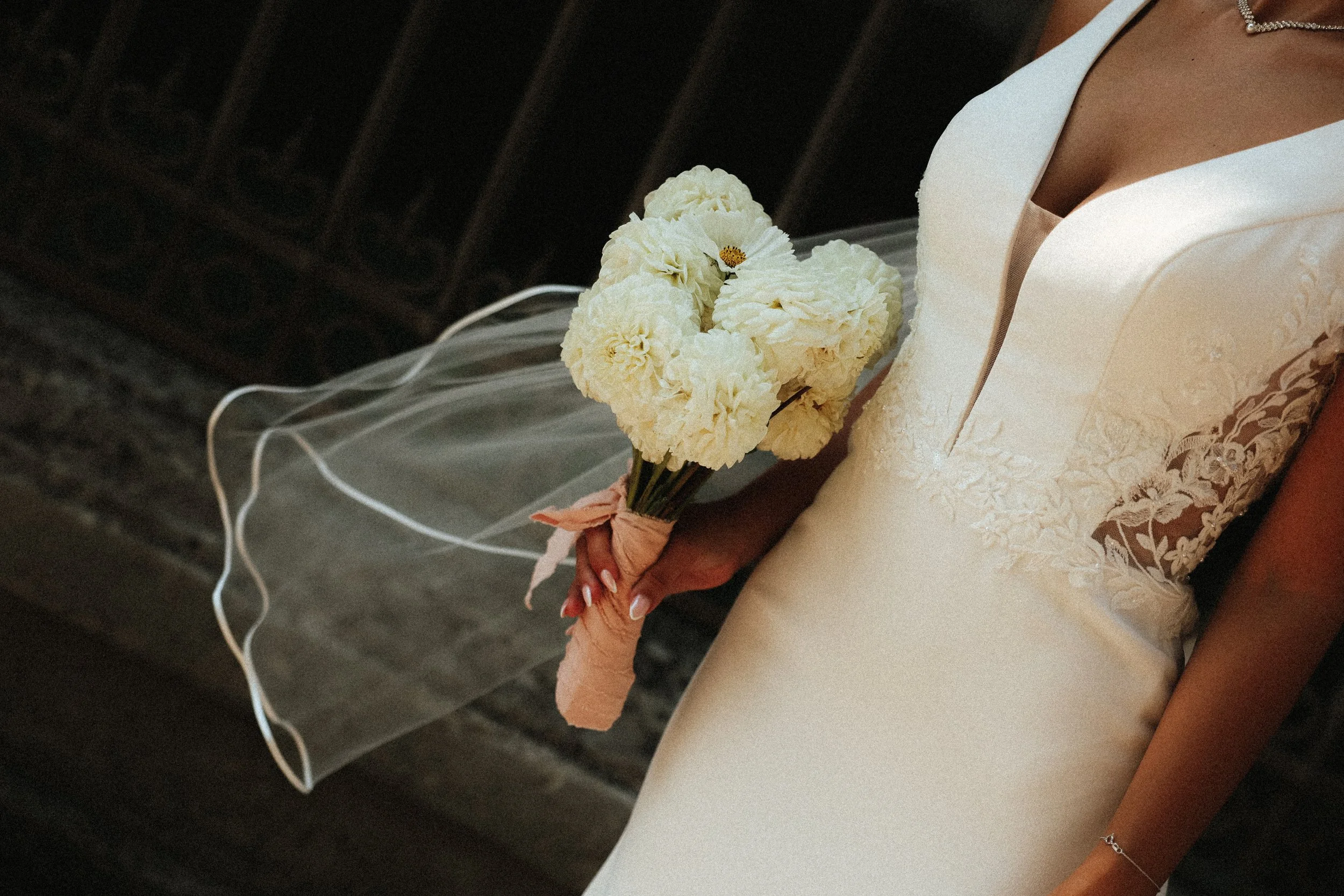 Une femme en robe de mariage ivoire tient un bouquet de fleurs blanches, comprenant des roses ou des dahlias, avec un ruban rose befesté autour du bouquet.