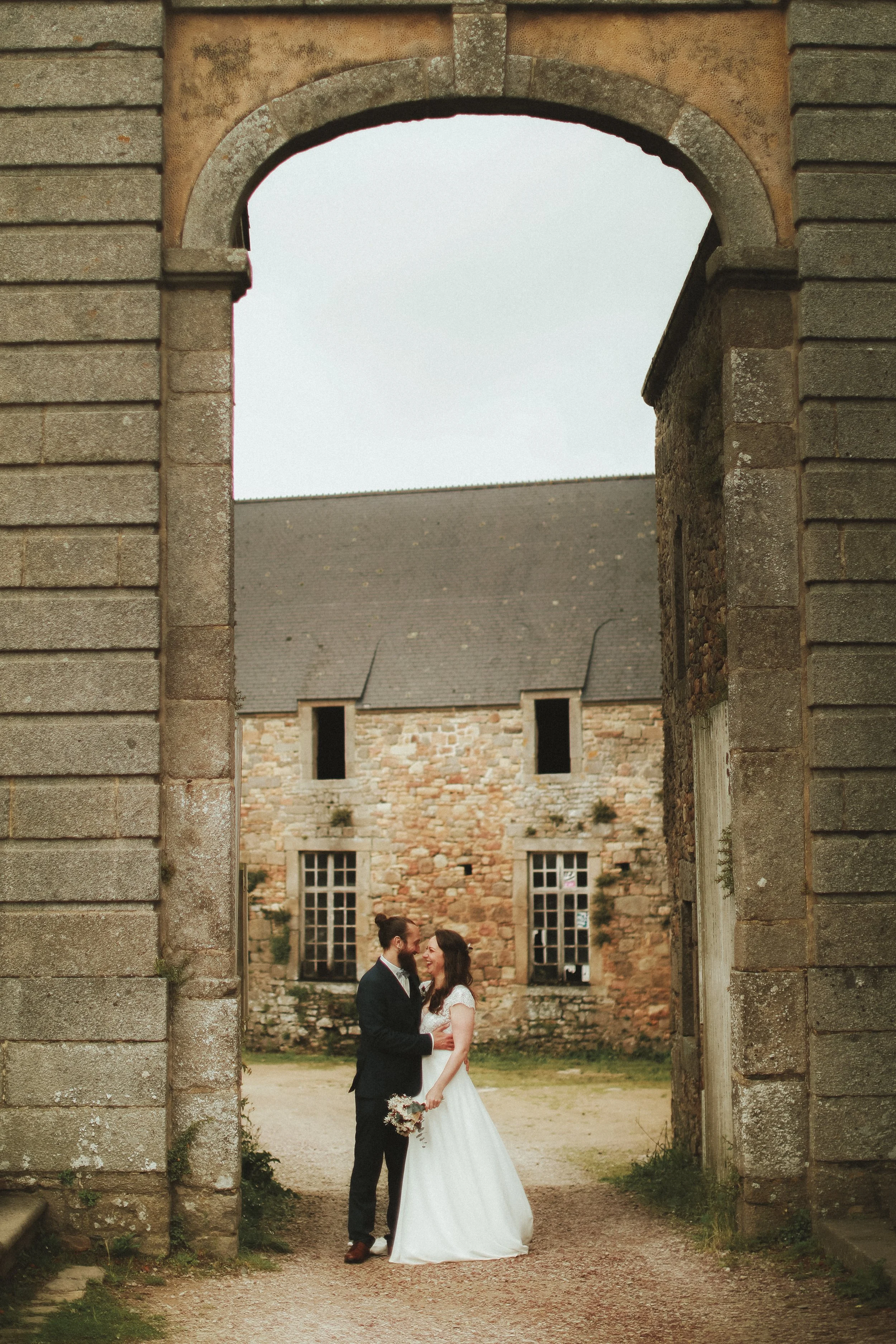 Un couple marié posant sous une arche en pierre devant un bâtiment ancien en pierre, avec des fenêtres en bois. La scène est en plein air, probablement dans un lieu historique ou un château, lors d'un mariage.