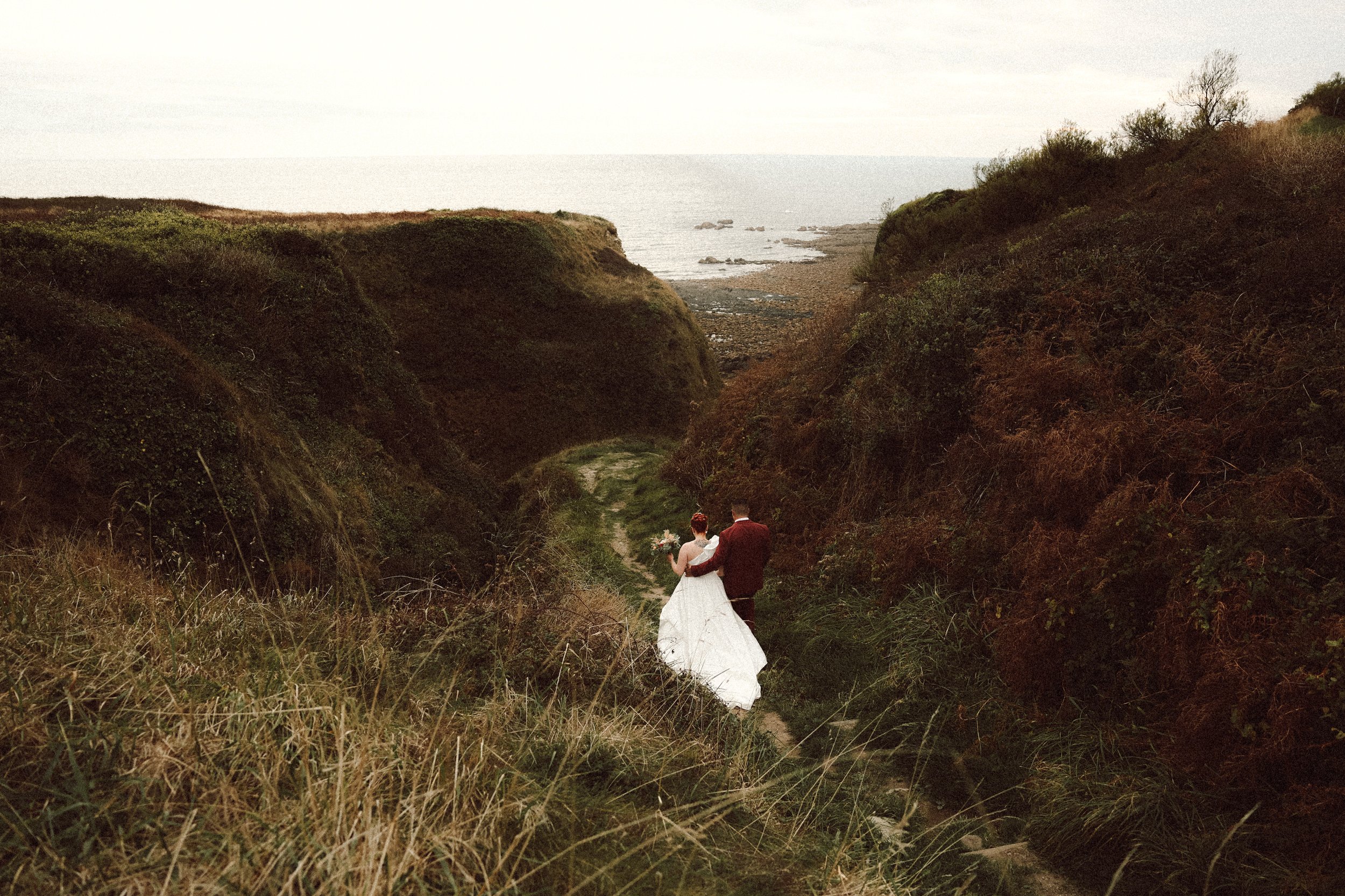 Un couple de mariés marchant dans un vallon verdoyant avec l'océan en arrière-plan, lors d'une séance photo de mariage.