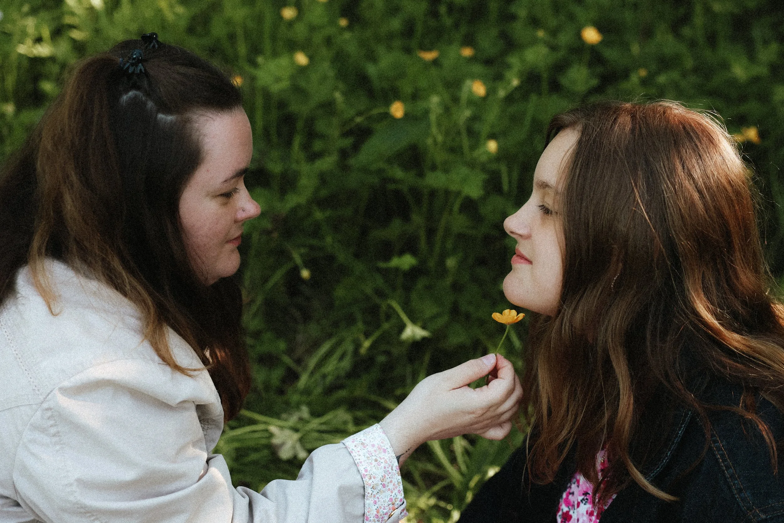 Deux jeunes femmes, l'une avec des cheveux bruns et l'autre avec des cheveux roux, échangent un regard dans un environnement naturel avec des fleurs et de la végétation.