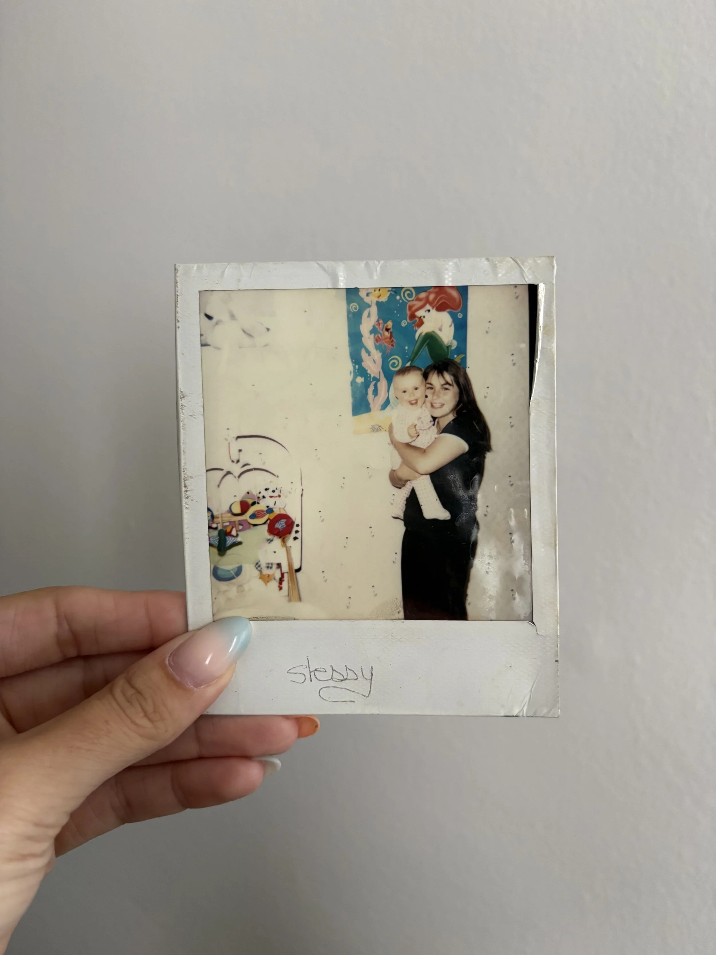 Une vieille photo polaroid montrant une femme et un bébé, avec une affiche de sirène et une œuvre d'art colorée en arrière-plan, la photo tient entre les doigts d'une main. La légende manuscrite en dessous indique 'stéphy'.