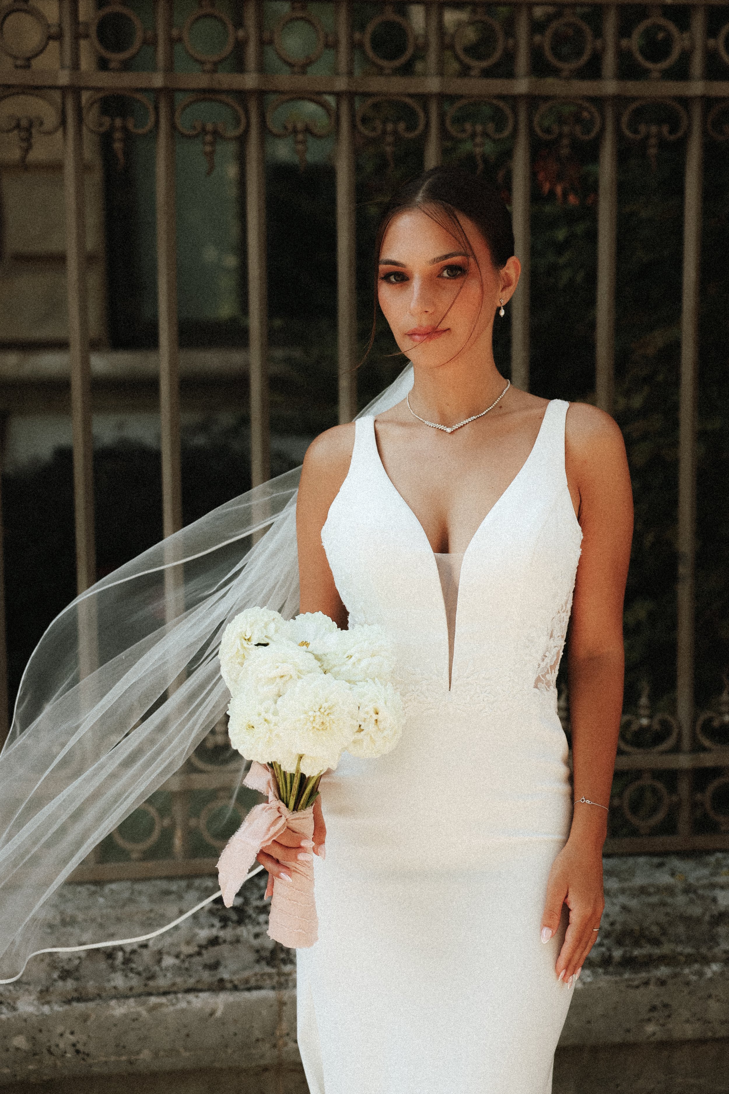 Une jeune femme en robe de mariée blanche tenant un bouquet de fleurs blanches, se tenant devant une grille en fer forgé.