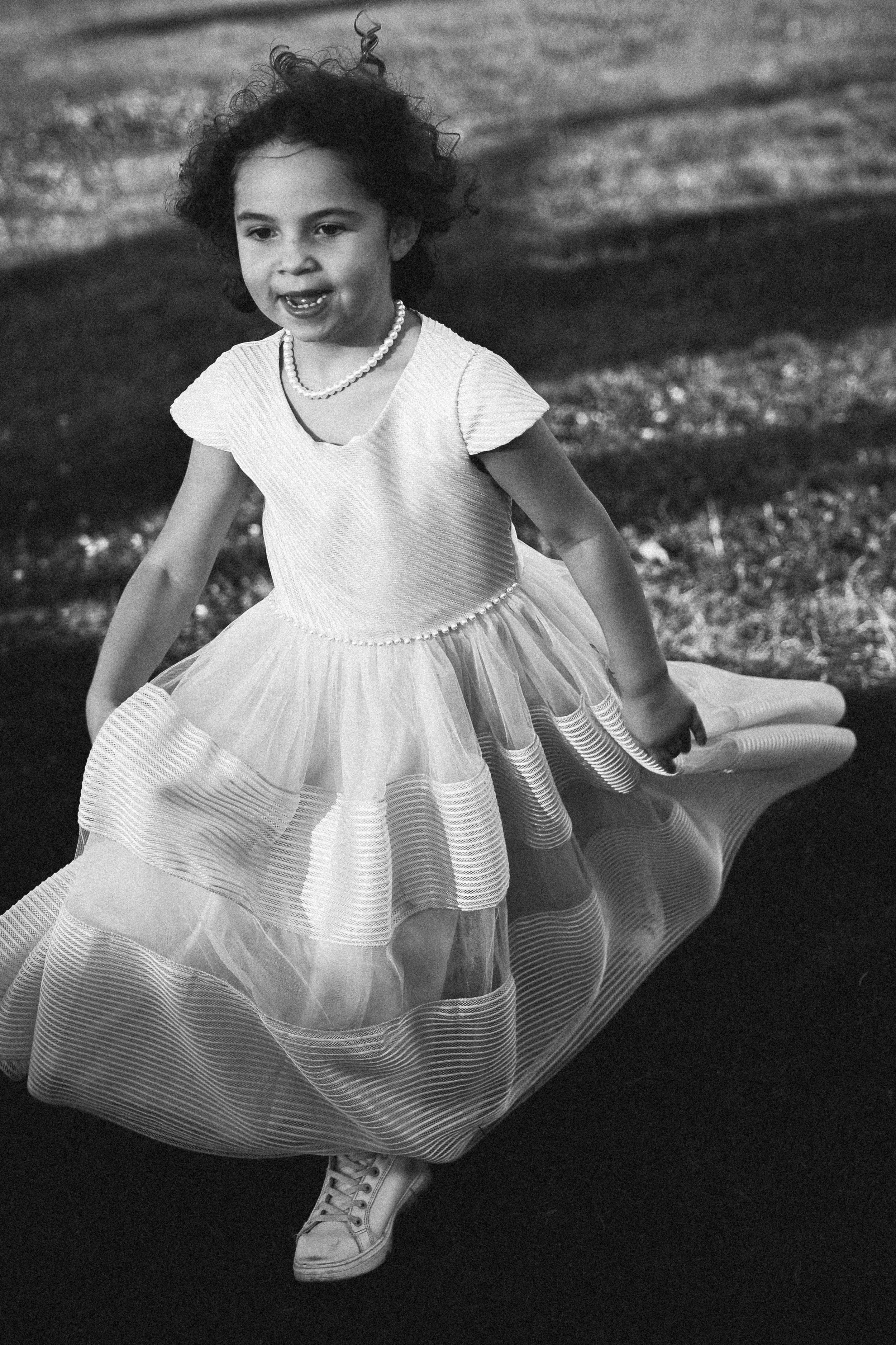 Une jeune fille qui court ou joue dans un espace extérieur, portant une robe élégante avec une jupe en tulle et des chaussures de sport, en noir et blanc.
