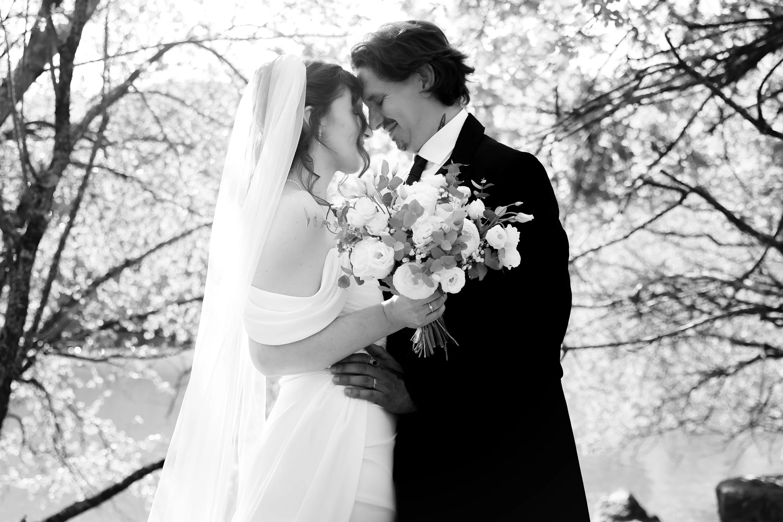 Un couple de mariés, une femme en robe de mariée avec un voile et un homme en costume noir, se rapproche avec leur front et leur nez en contact. Elle tient un bouquet de fleurs blanches et vertes, et ils sont sous des arbres, en plein air.
