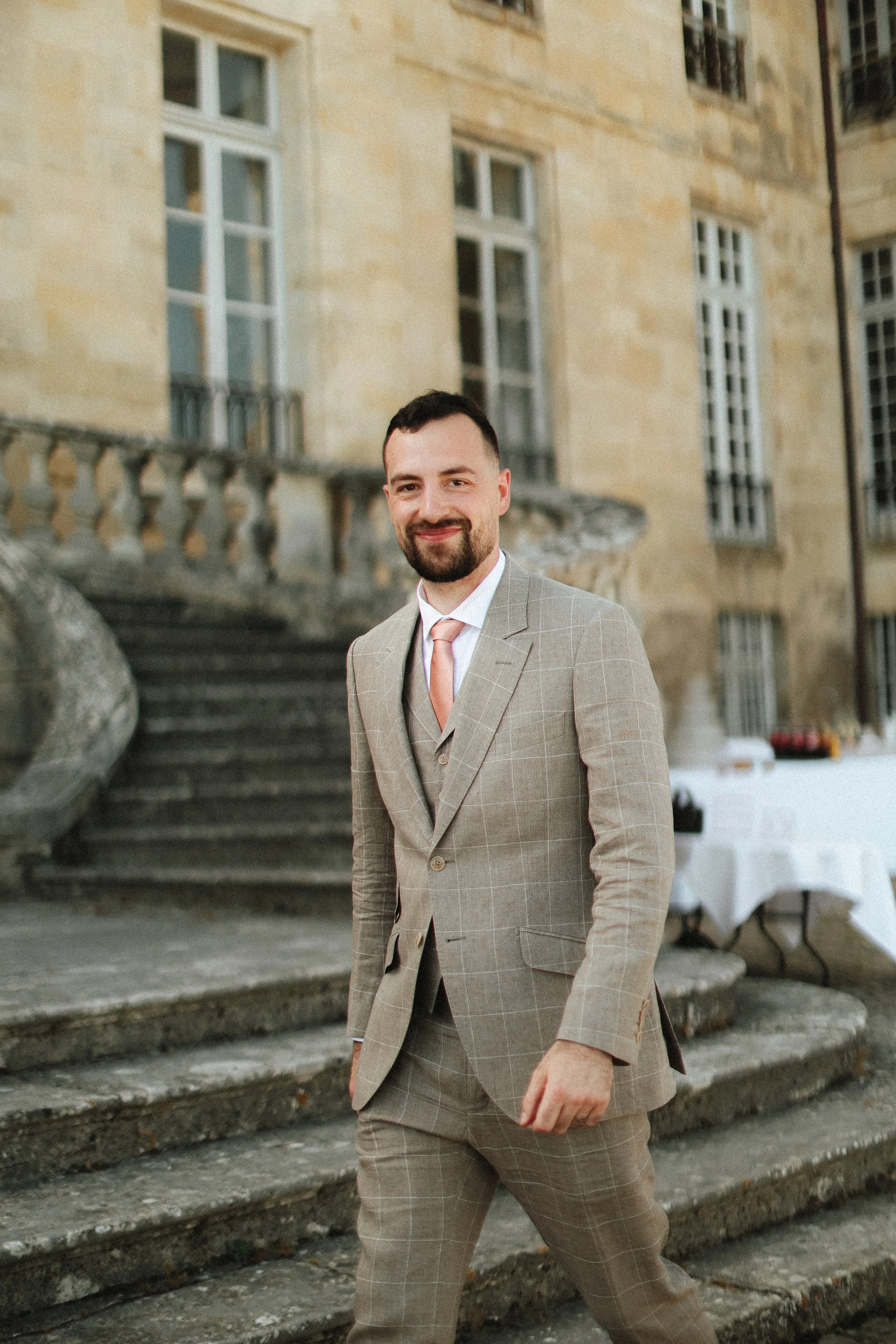 Un homme souriant en costume beige à carreaux, avec une cravate rose, en extérieur devant un bâtiment ancien en pierre, près de marches en pierre.
