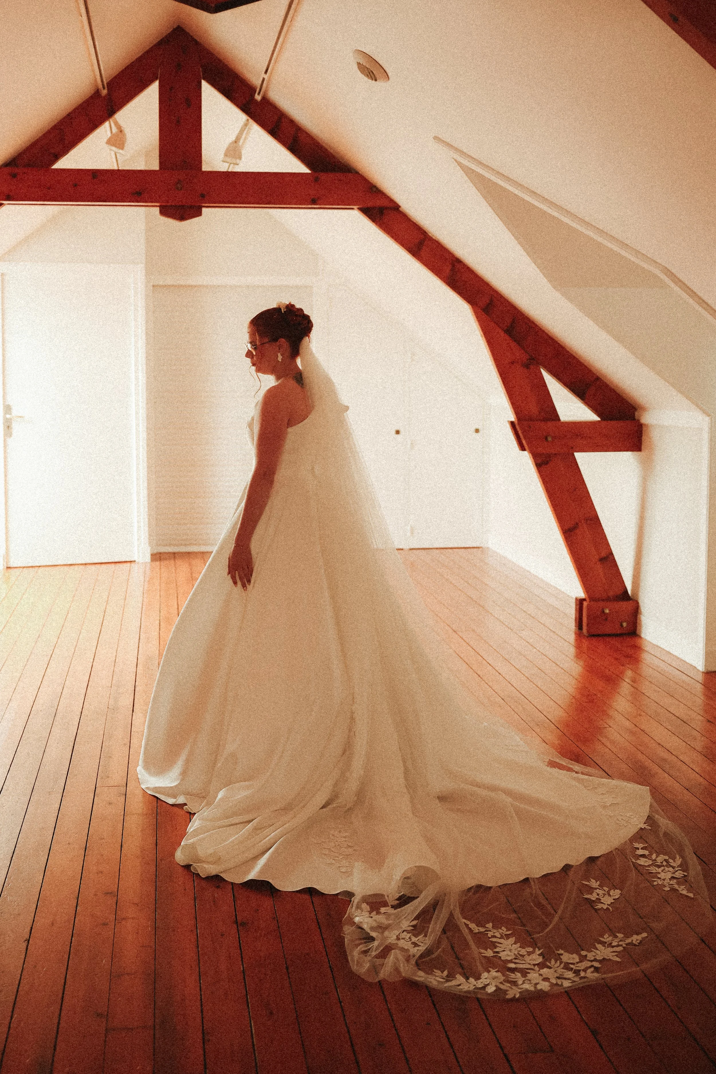 Une femme en robe de mariage dans une pièce avec plafond en pente et poutres en bois, éclairée par la lumière naturelle