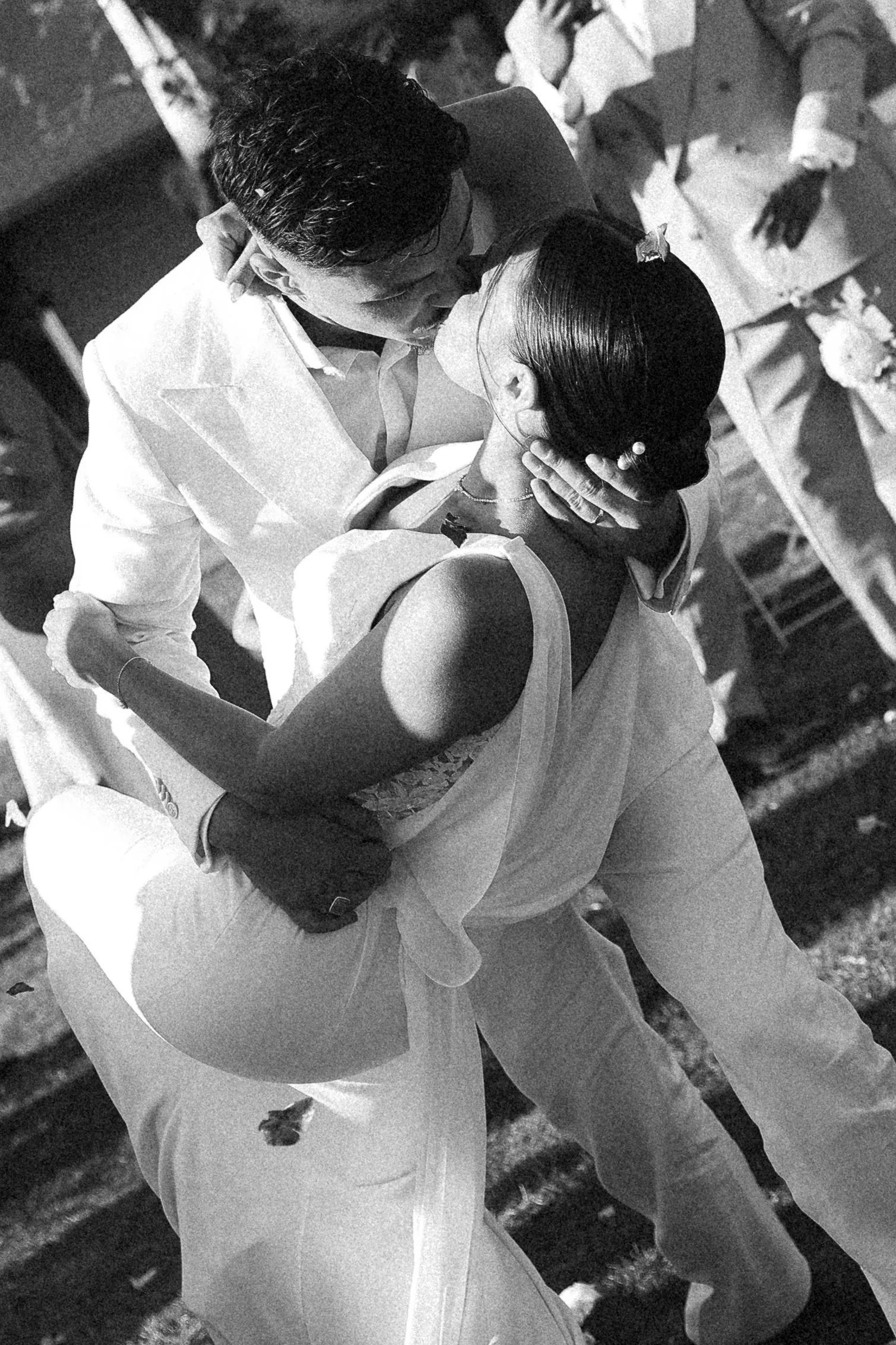 Un couple danse lors d'une cérémonie en noir et blanc, l'homme porte un costume blanc et la femme une robe blanche avec des épaules dénudées, ils s'embrassent tendrement.