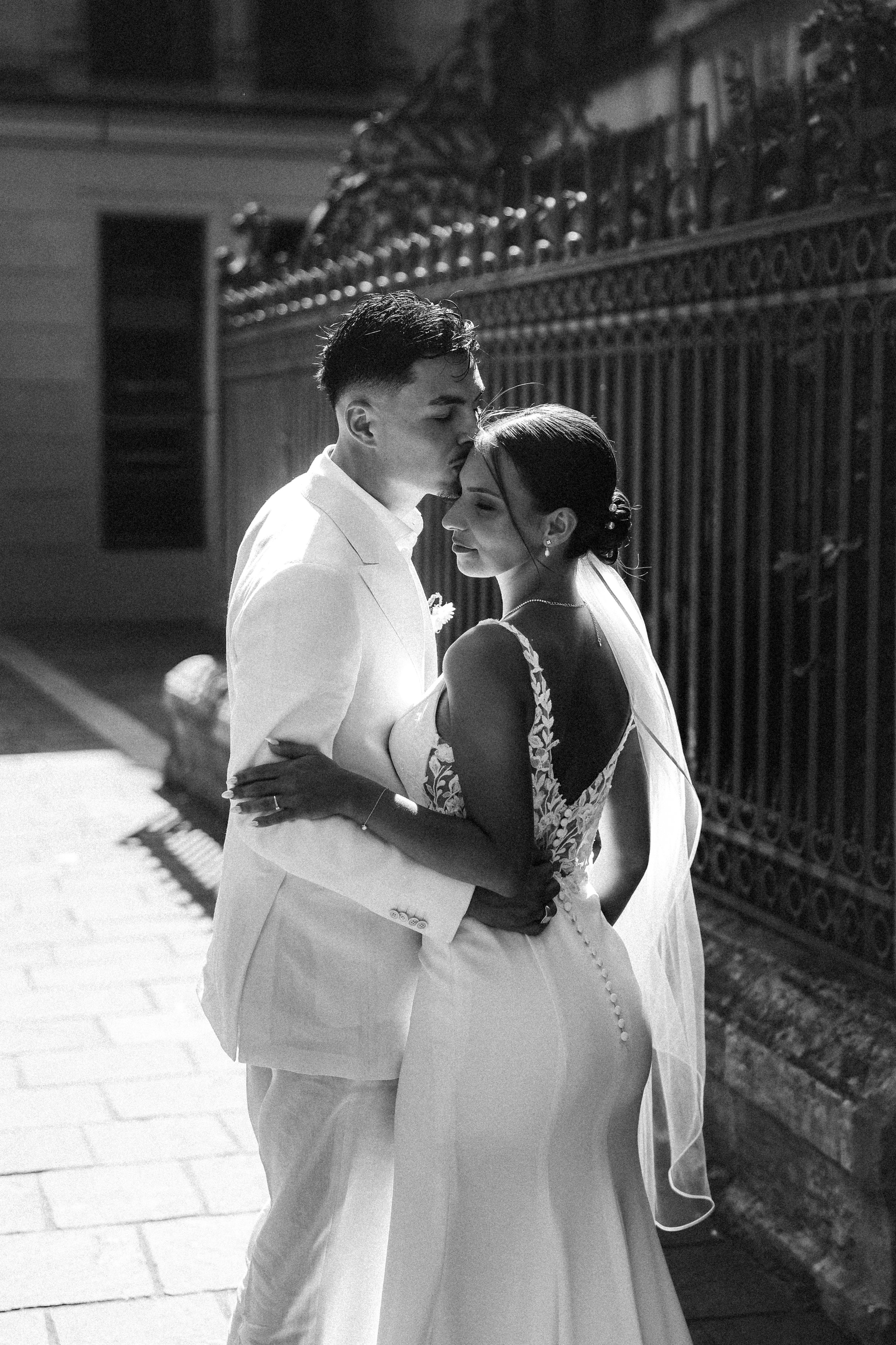 Un couple de mariés en habits de mariage pose tendrement devant une grille en métal noir lors de la nuit. L'homme en costume blanc embrasse la femme sur le front.