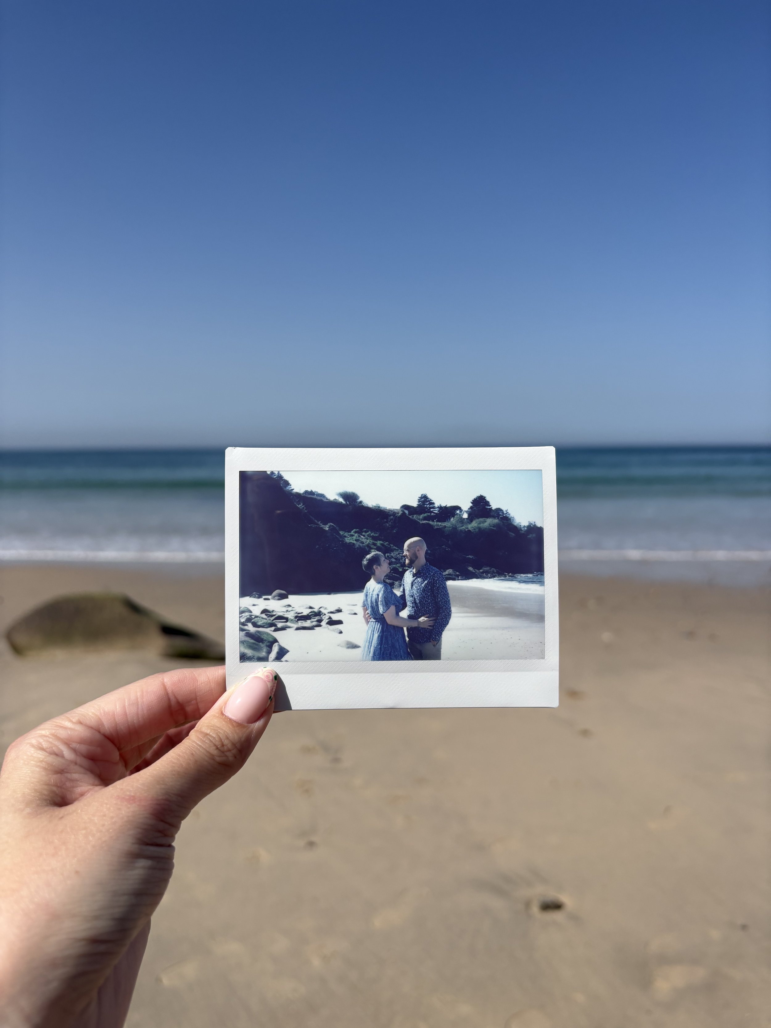 Une personne tient une photo instantanée d'un couple regardant dans les yeux sur une plage, avec la mer et le ciel bleu en arrière-plan.