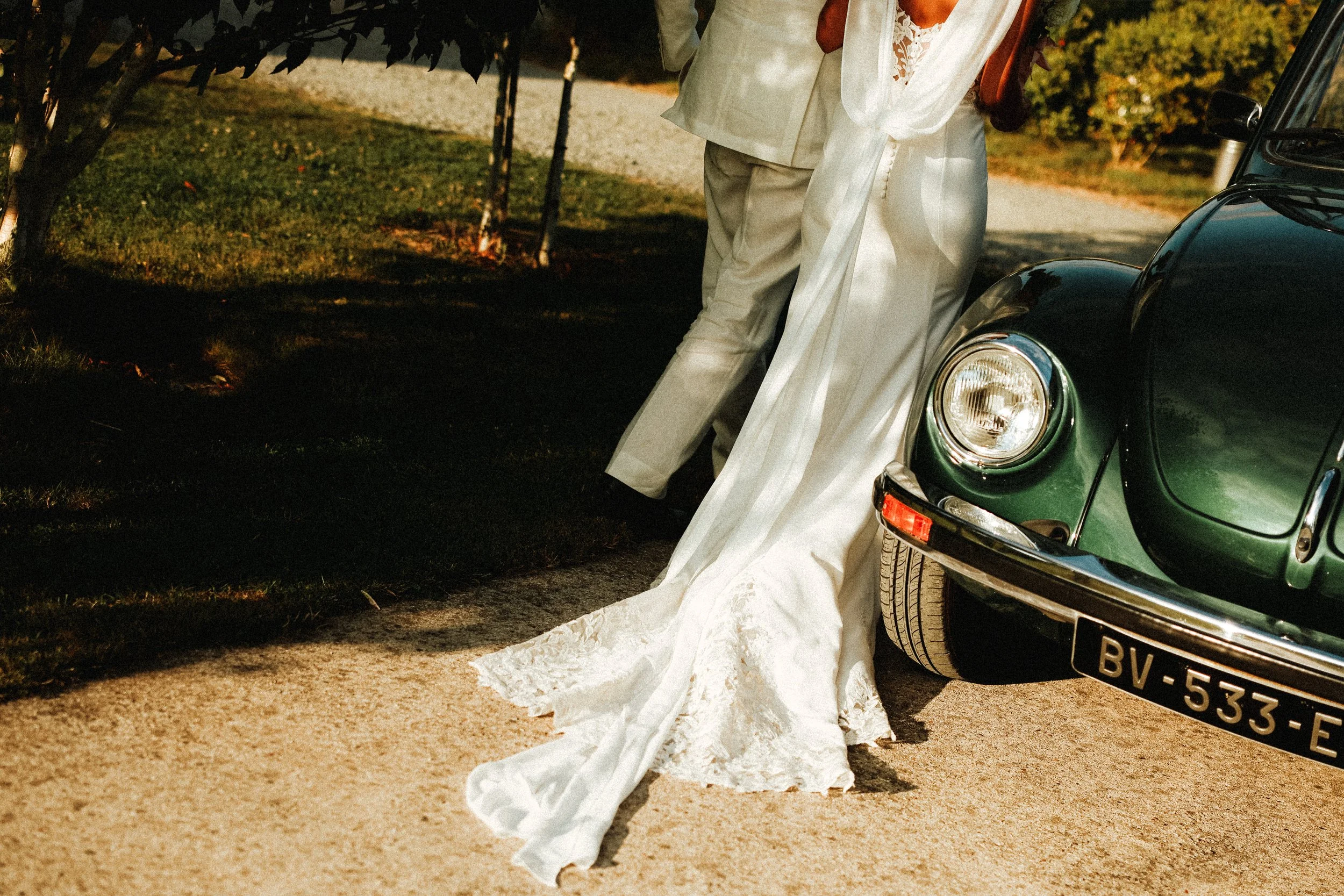 Un couple en vêtements de mariage posant près d'une voiture ancienne verte, avec une robe de mariée en satin blanc et un costume beige, sur un chemin pavé entouré d'arbres en automne.