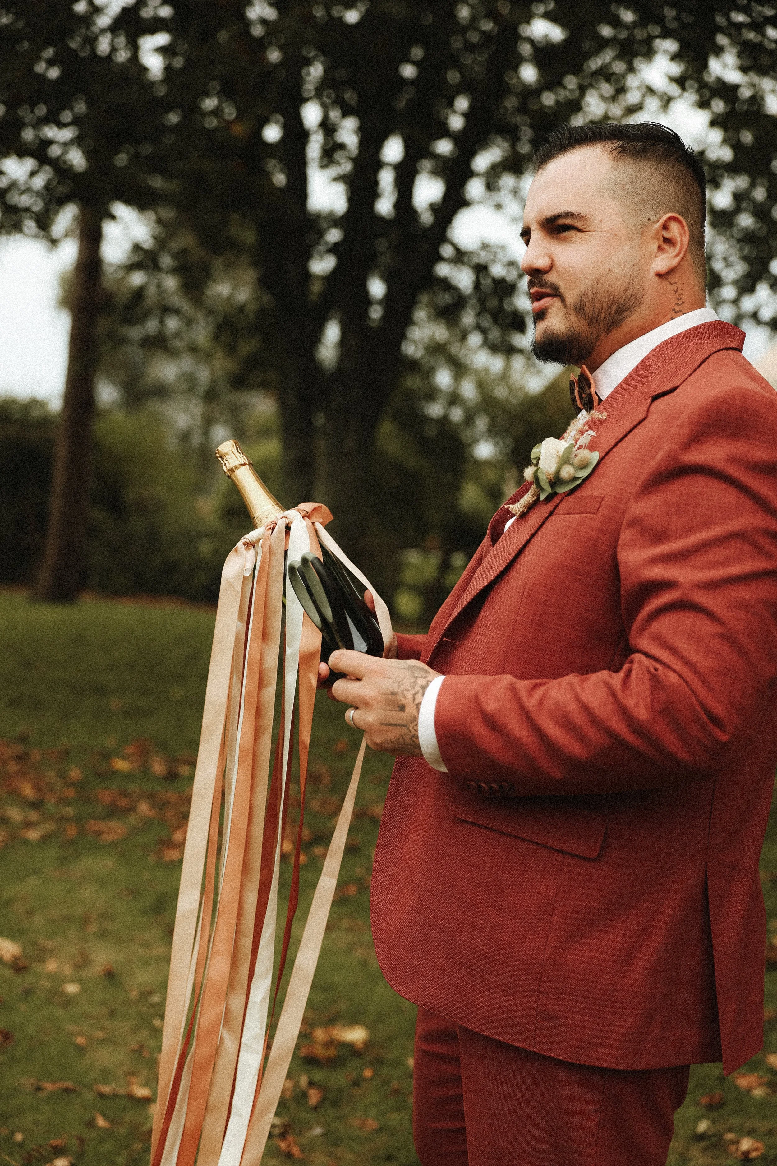 Un homme en costume rouge tient une bouteille de champagne décorée de rubans et vérifie son téléphone lors d'un mariage en extérieur.