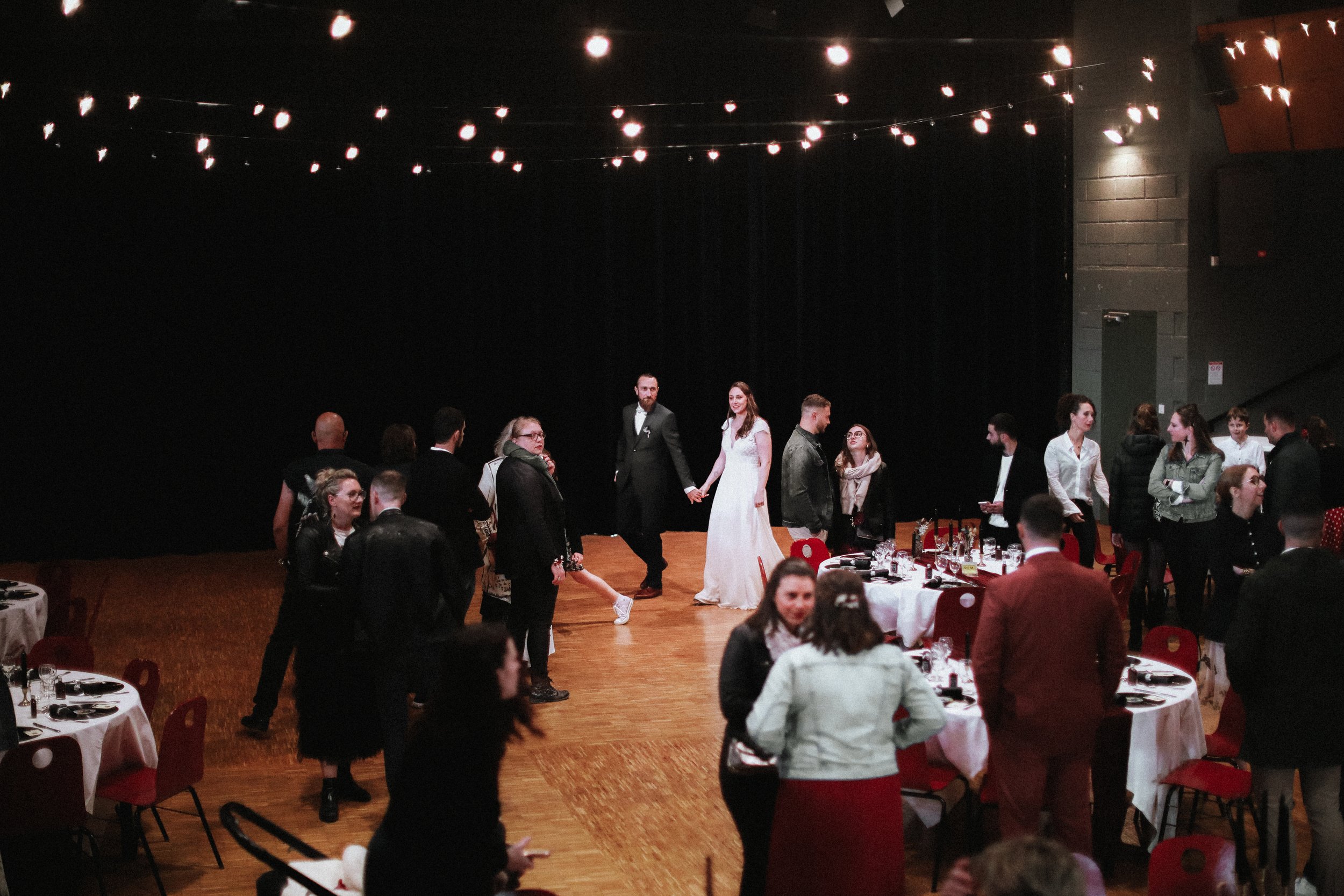 Un groupe de personnes lors d'une réception ou d'un mariage, certaines dansant au centre de la salle, d'autres autour des tables, décorée avec des lumières suspendues au plafond.