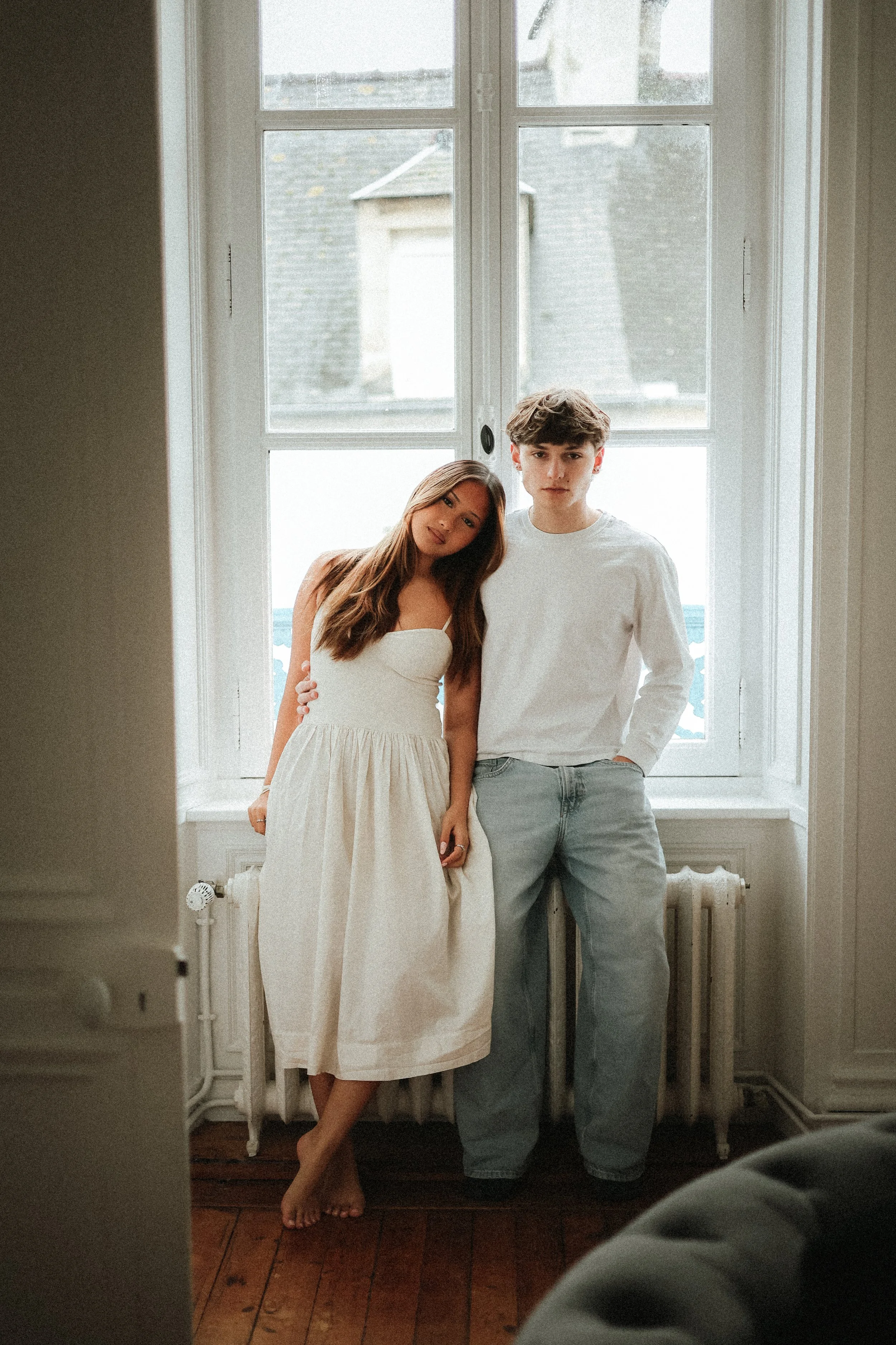 Deux jeunes debout près d'une grande fenêtre, l'un porte une robe blanche et l'autre un t-shirt blanc et un jeans, dans une pièce lumineuse avec un parquet en bois.