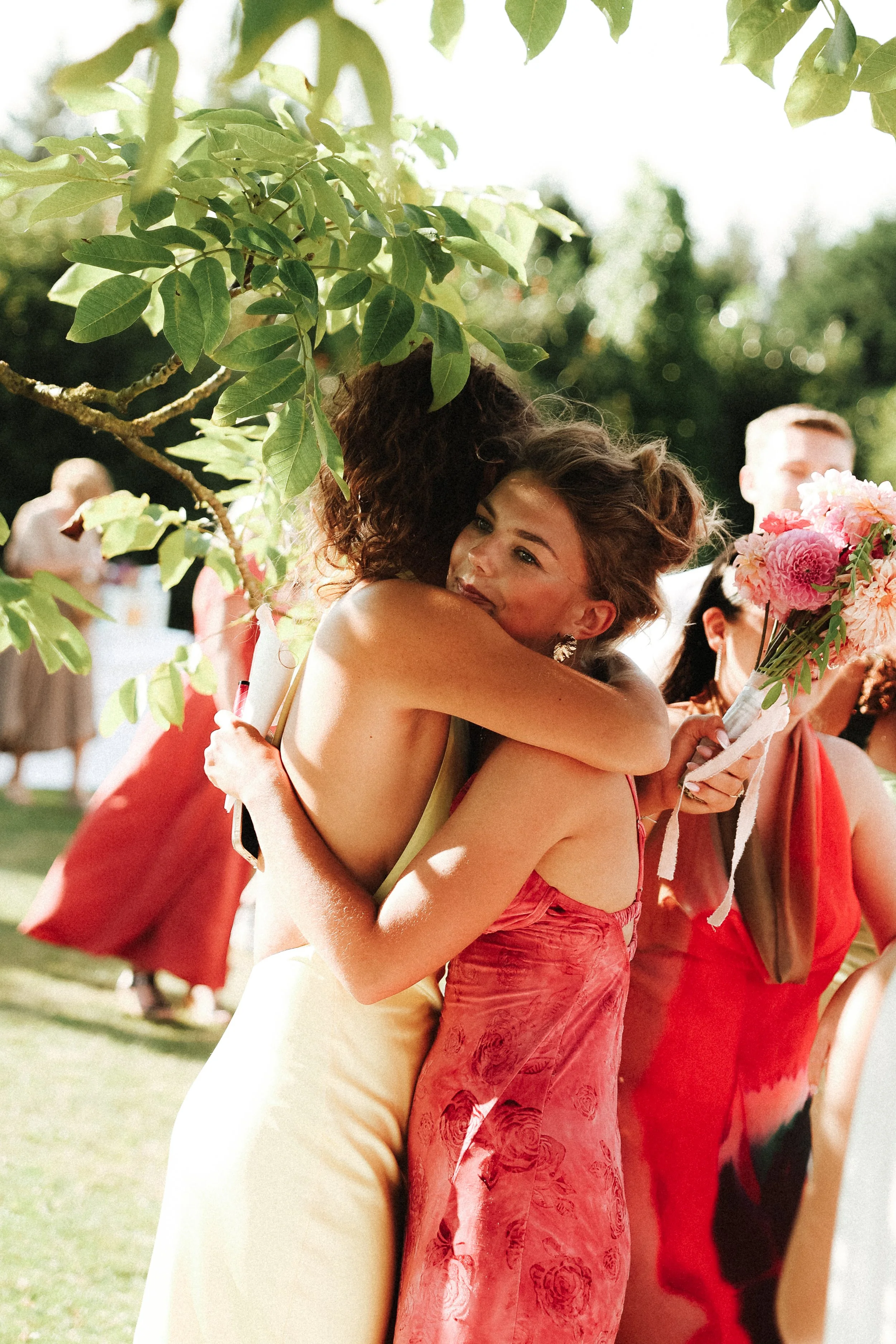 Deux femmes en robes d'été s'embrassent lors d'une célébration en extérieur, entourées d'autres personnes.