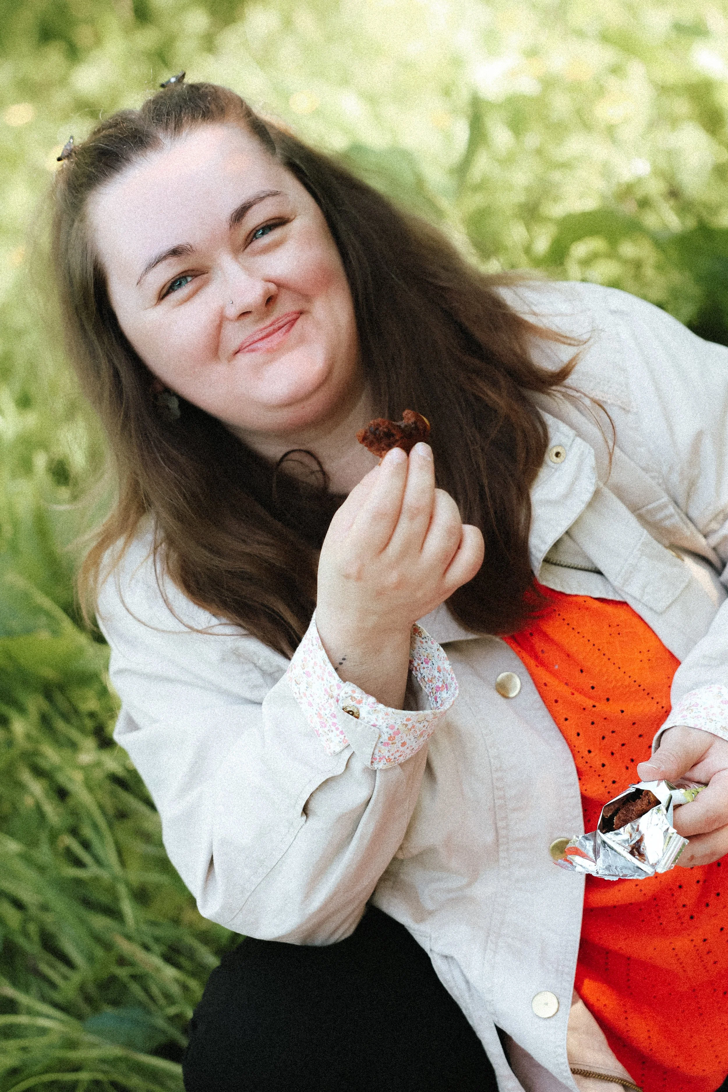 Une jeune femme avec des cheveux bruns, portant une veste beige, une chemise à manches longues à motifs et un pantalon noir, souriant en tenant un morceau de gâteau dans une main et une barre de chocolat dans l'autre. Elle est dans un environnement v