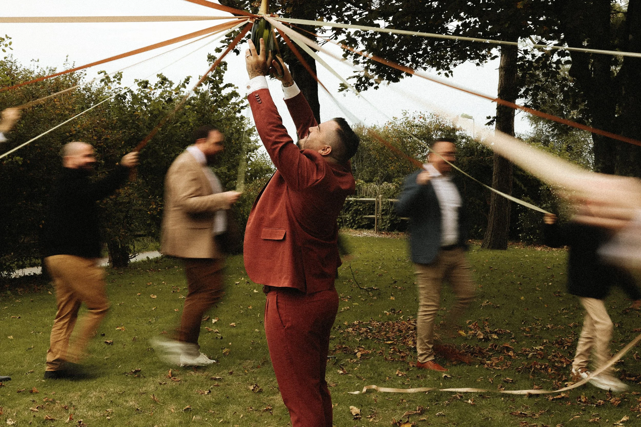 Groupe de personnes en costume, dansant et jouant à la tapette à souris en extérieur dans un parc avec des arbres, sous un ciel nuageux, lors d'un événement ou fête.