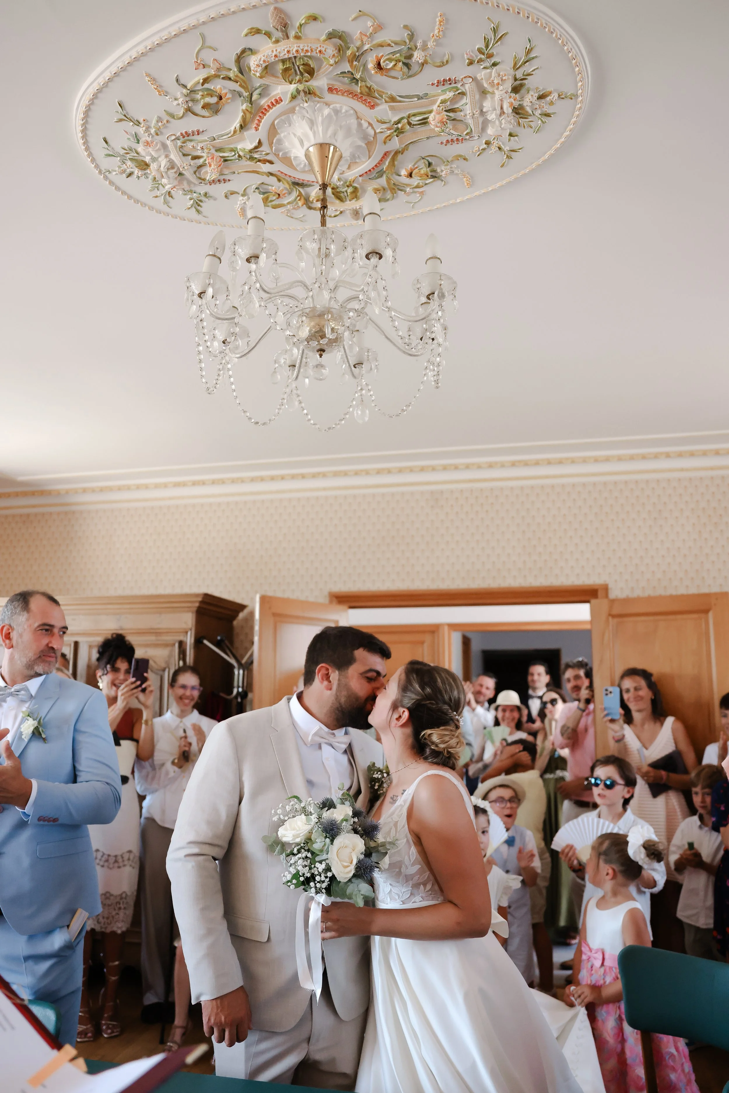 Un couple de mariés s'embrassant lors de leur cérémonie dans une pièce décorée élégamment, entourés de témoins et d'invités qui prennent des photos.