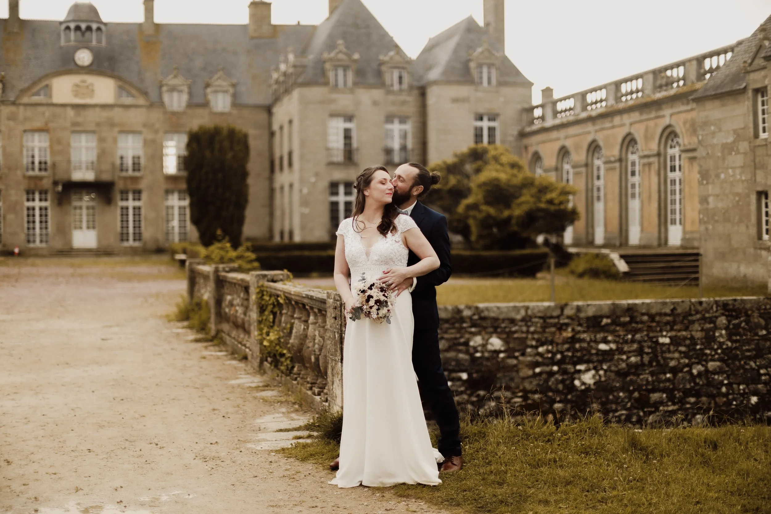 Un couple de mariés posant devant un château historique, l'homme embrassant tendrement la femme qui tient un bouquet de fleurs, lors d'une séance de photographie nuptiale.