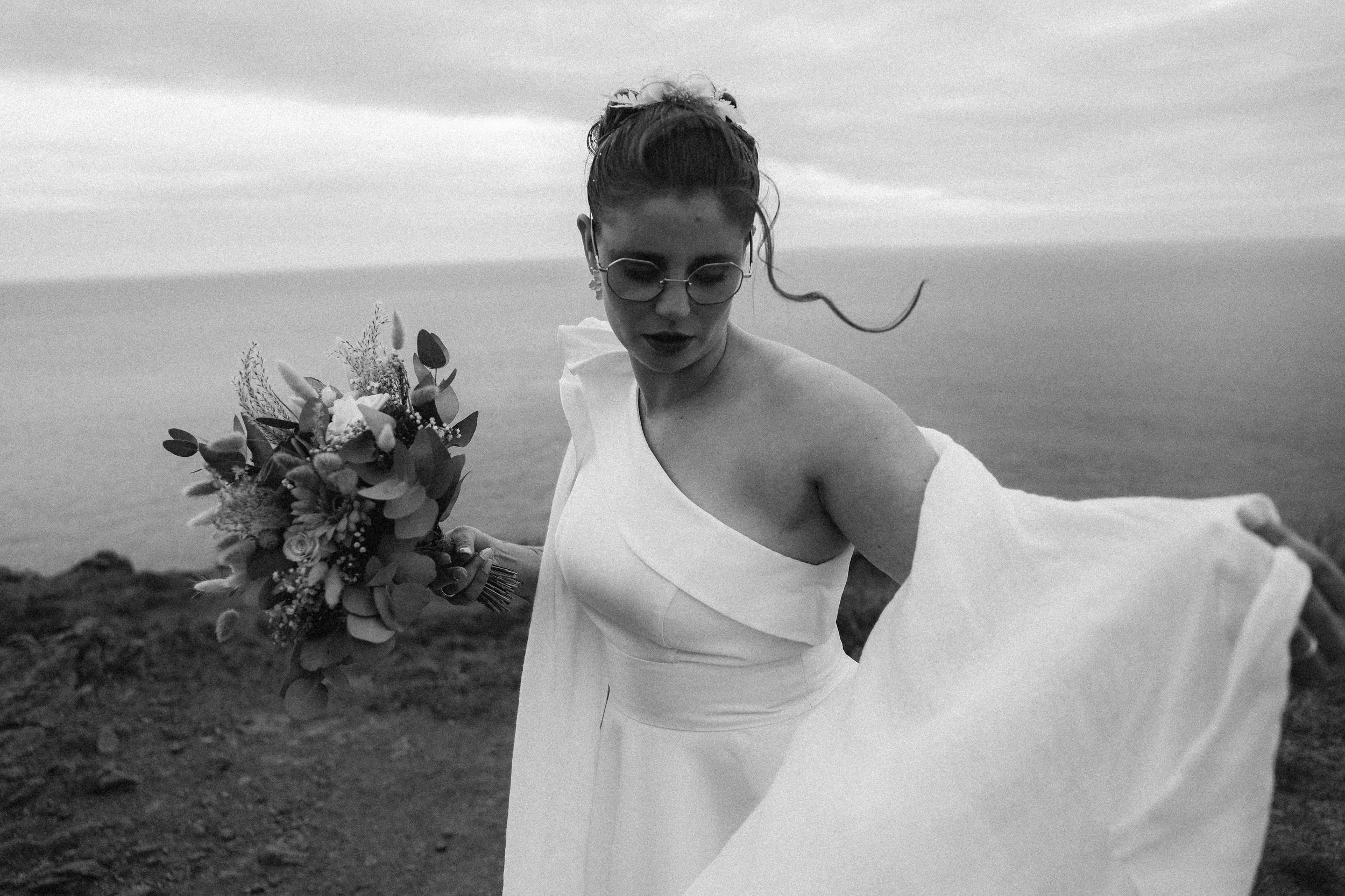 Une femme portant une robe blanche et des lunettes de soleil, tenant un bouquet de fleurs, se tenant sur une falaise avec vue sur la mer. Elle regarde vers l'objectif, avec une expression mystérieuse.