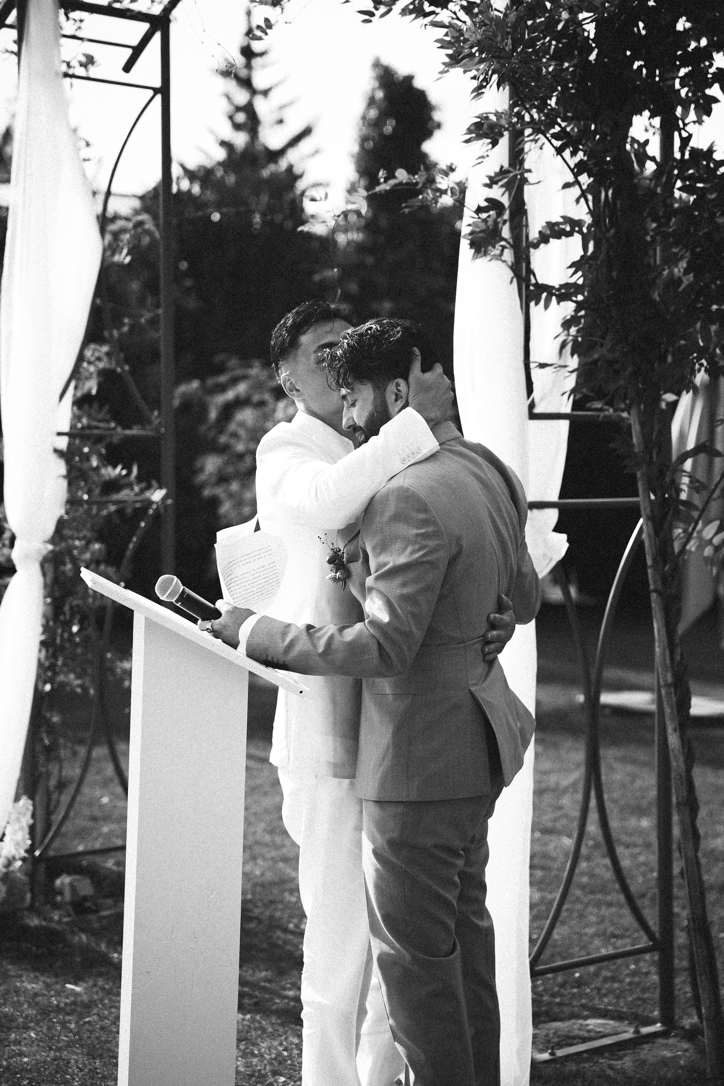 Deux hommes, un en costume blanc et l'autre en costume gris, enlaçant lors d'une cérémonie en plein air, avec un micro et un pupitre.