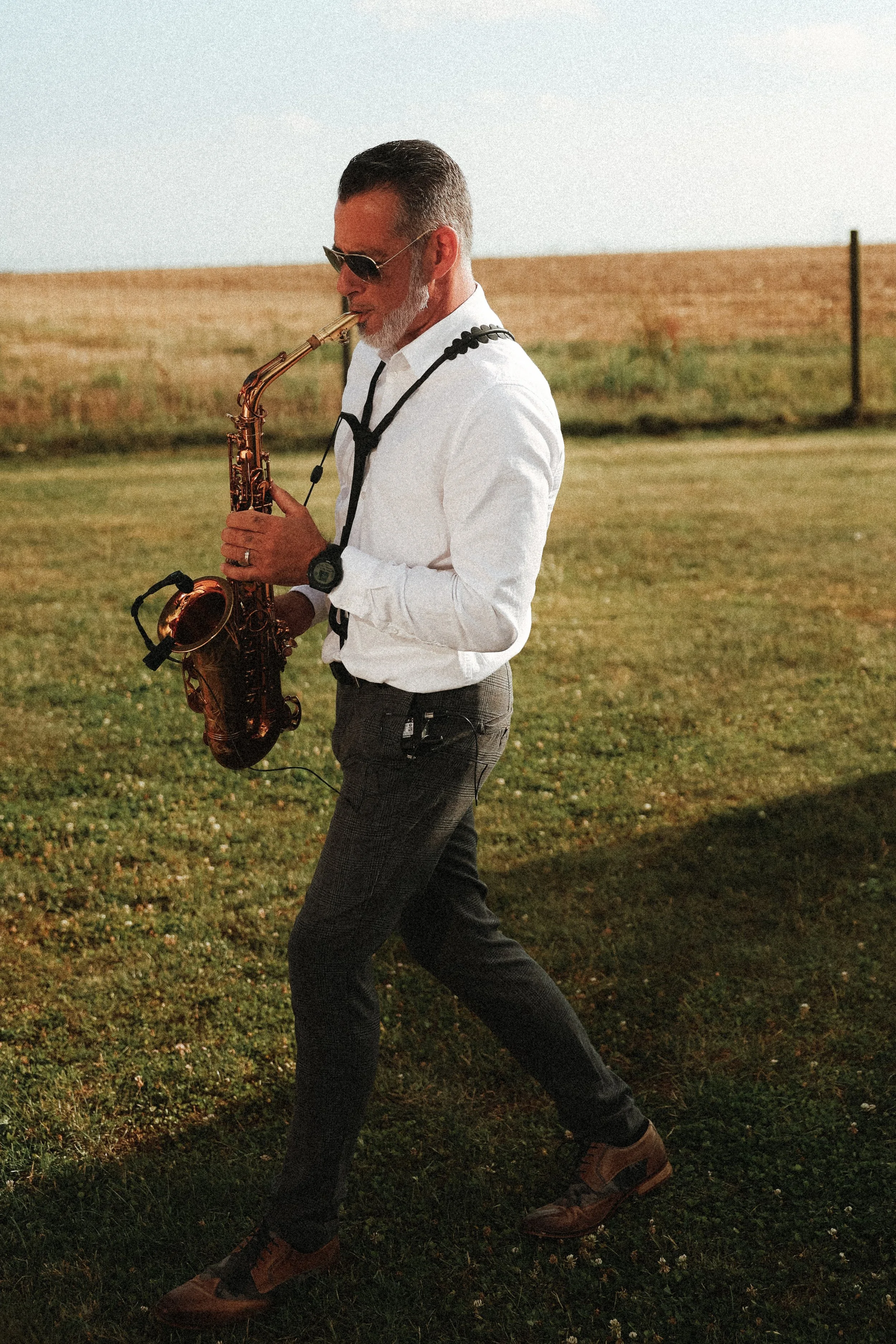 Homme jouant du saxophone en plein air, vêtu d'une chemise blanche, pantalon sombre et lunettes de soleil, dans un paysage rural avec un champ en arrière-plan.