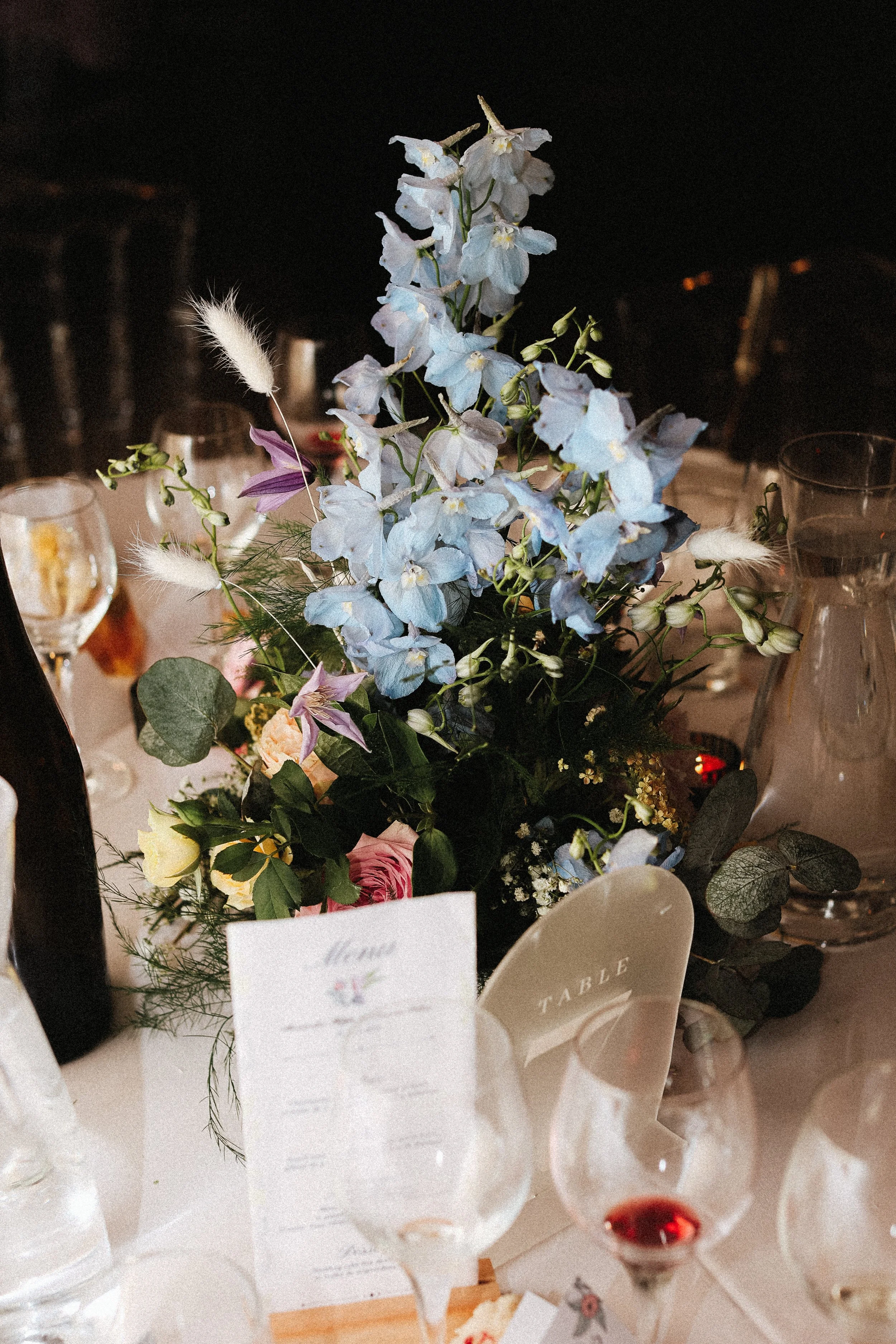 Une composition florale avec des fleurs bleues, violettes, jaunes et blanches sur une table de dîner décorée, avec des verres, une carte de menu et une carafe d'eau.