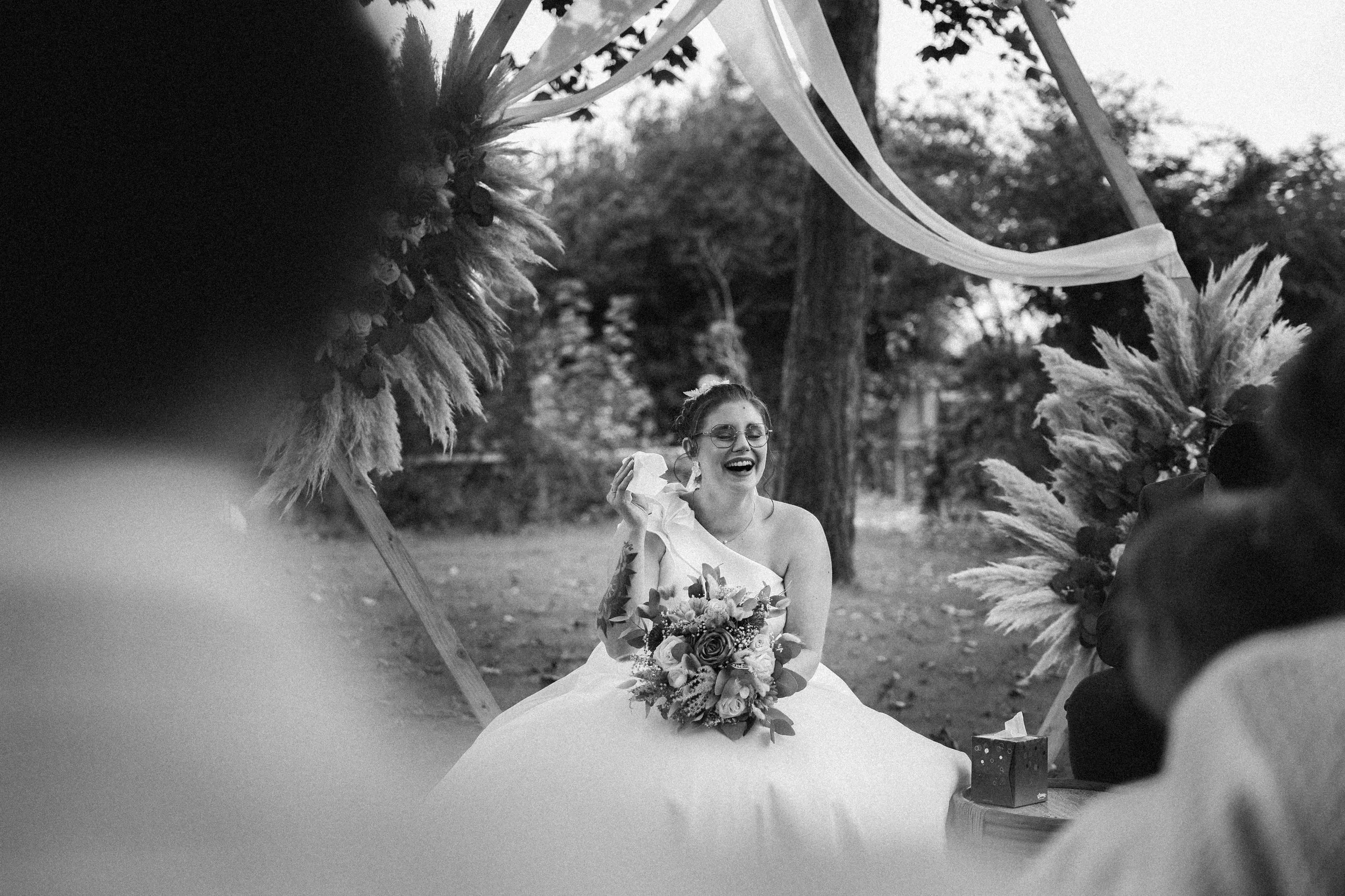 Une mariée souriante portant une bouquet de fleurs, assise en plein air, entourée de décorations de mariage en plumes, avec des arbres en arrière-plan. Sketch en noir et blanc.