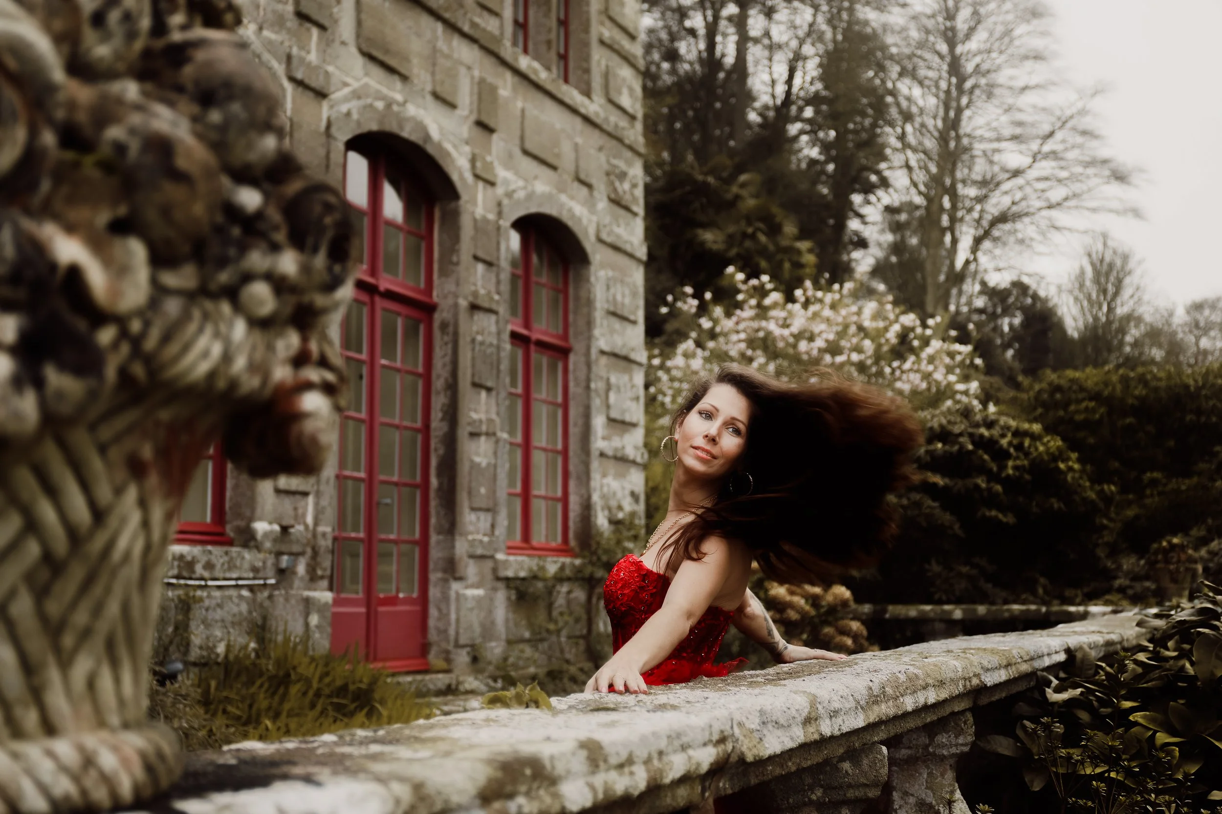 Une femme en robe rouge regarde vers la caméra en posant sur un muret en pierre devant une vieille maison en pierre avec des fenêtres à cadre rouge et un jardin végétalisé en arrière-plan, avec des arbres et des fleurs.