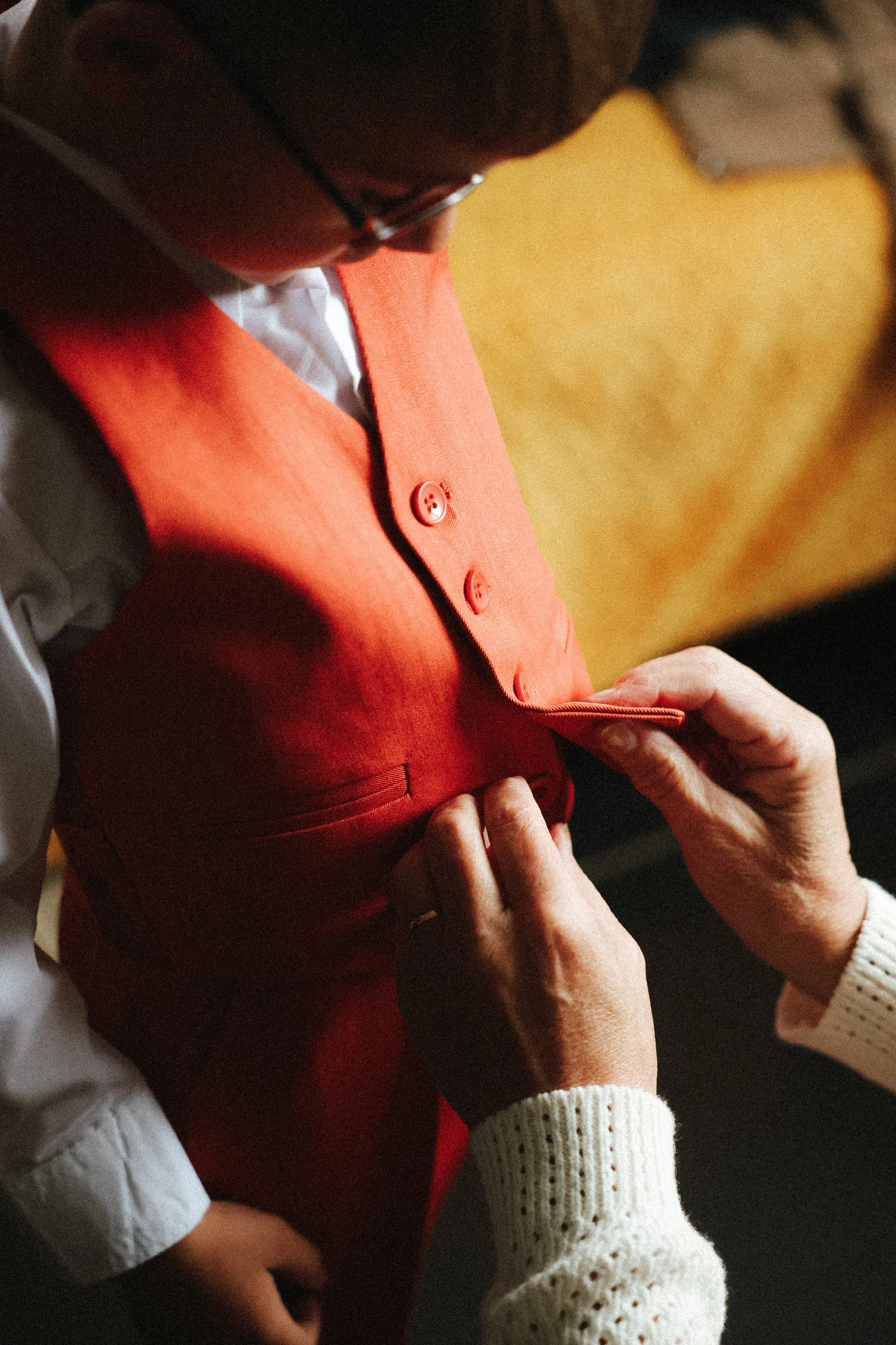 Une personne aide à boutonner un vêtement orange porté par une autre personne, une femme âgée. La scène se déroule en intérieur avec un fond beige.