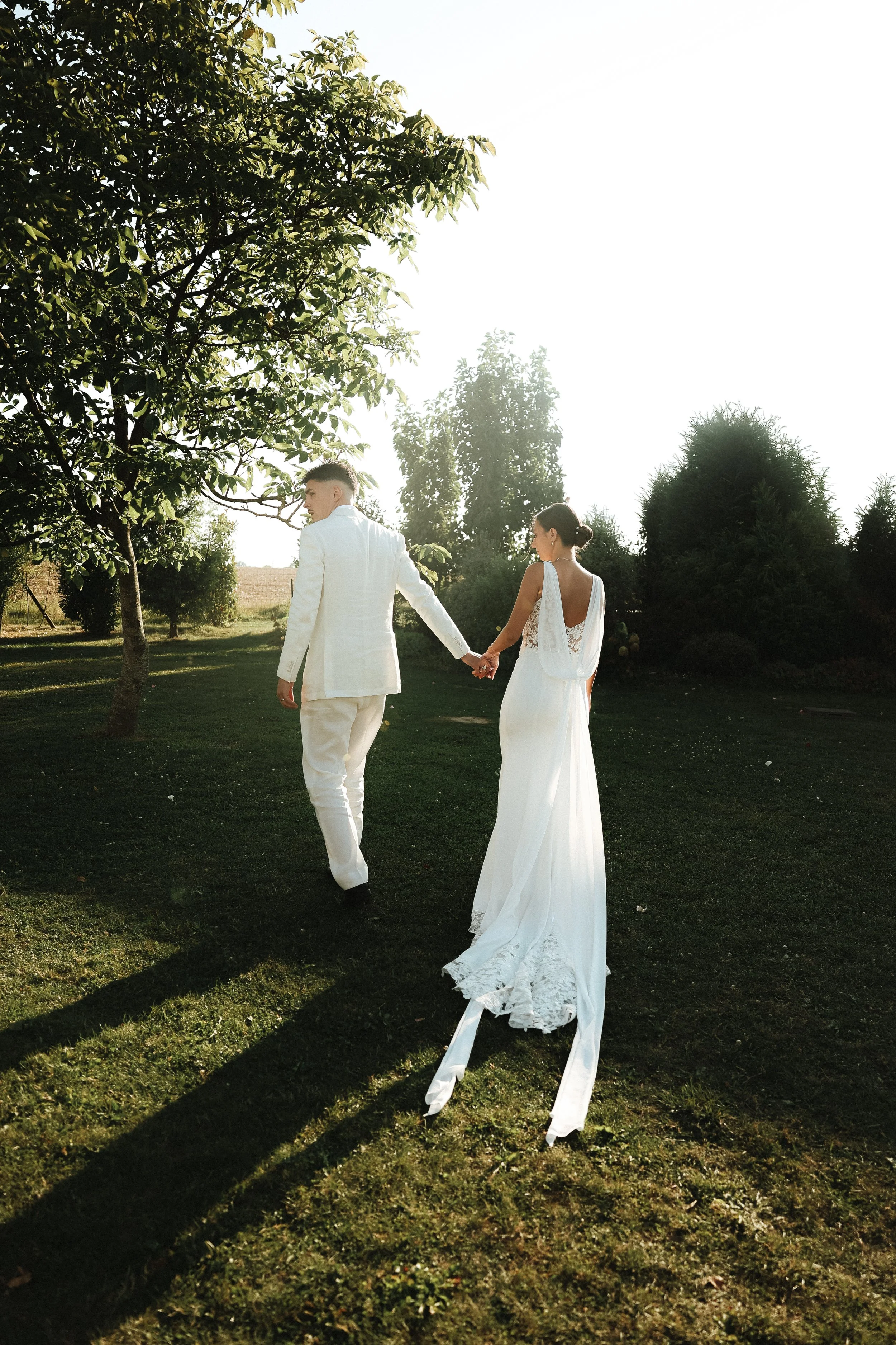 Un couple en tenue de mariage marche dans un jardin ensoleillé, se tenant la main, avec des arbres en arrière-plan.