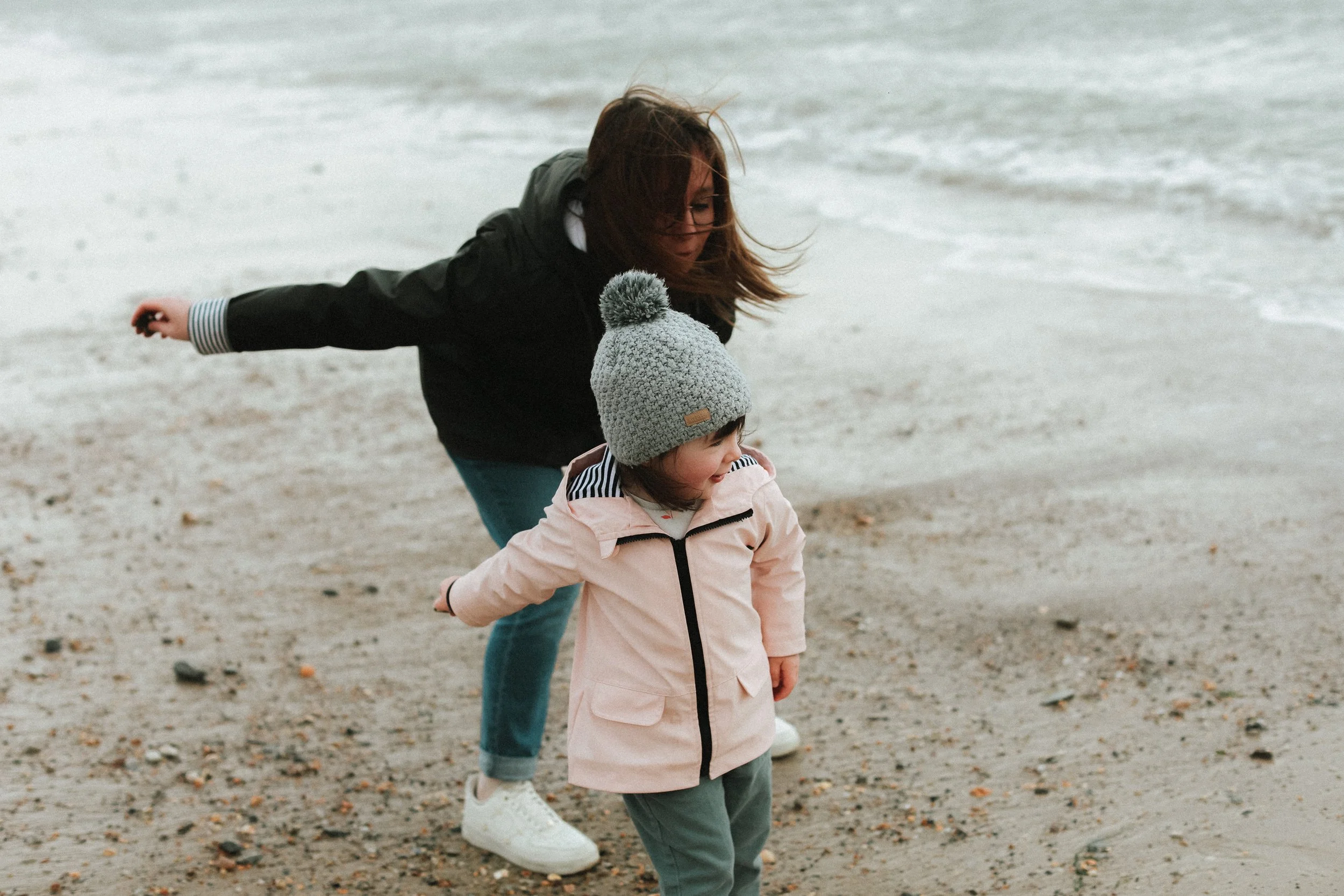Une femme et une petite fille jouent près de la mer sur la plage. La femme, avec des cheveux bruns, porte une veste noire, tandis que la petite fille porte un bonnet gris, un manteau rose et un pantalon gris. Le vent souffle et il fait probablement f