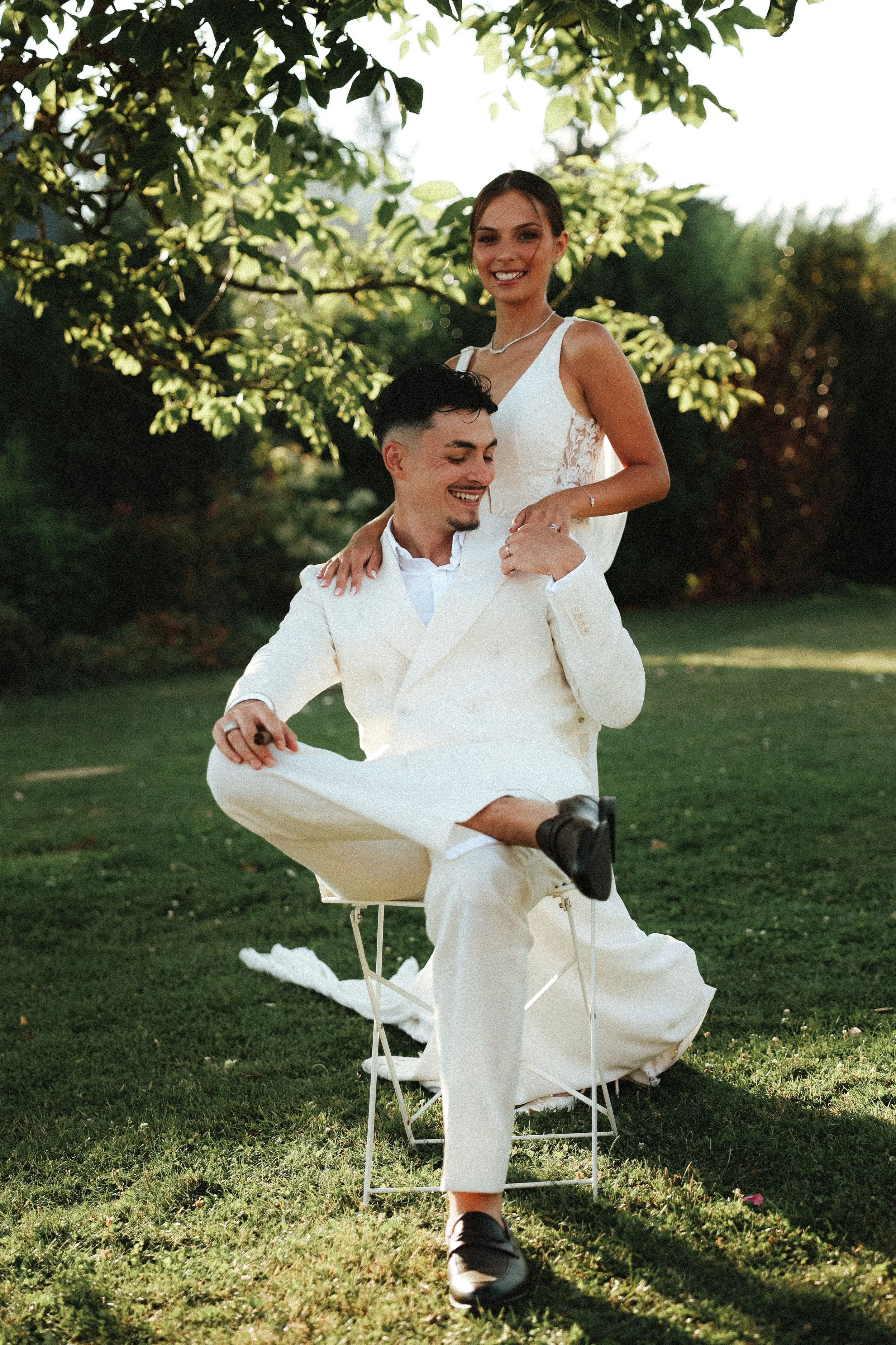 Un couple en tenue de mariage dans un jardin, l'homme assis sur une chaise en train de rire et la femme debout derrière lui, souriante.
