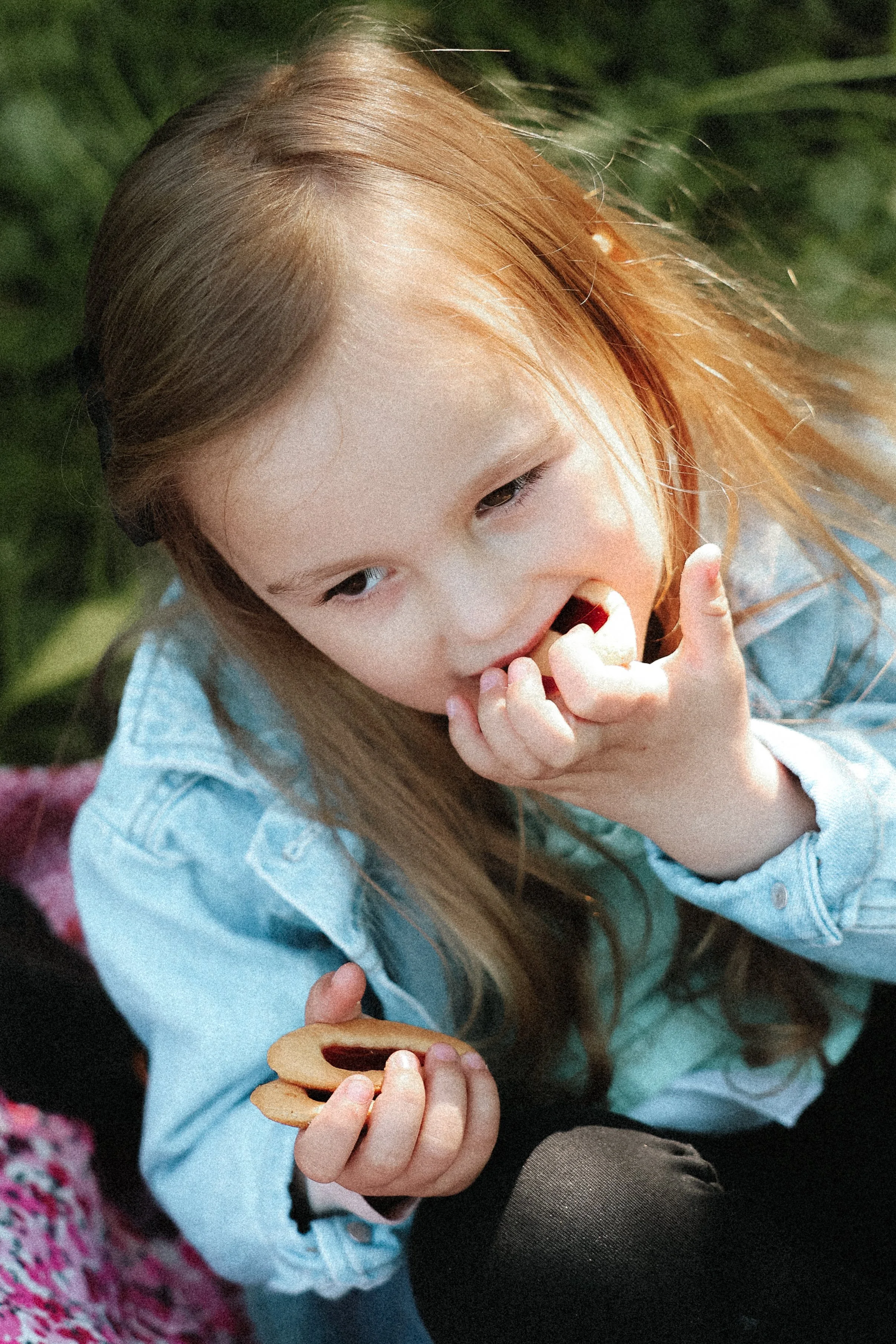 Une jeune fille aux cheveux roux qui sourit et mange un biscuit, assise sur un tapis avec un autre enfant, dans un environnement extérieur.