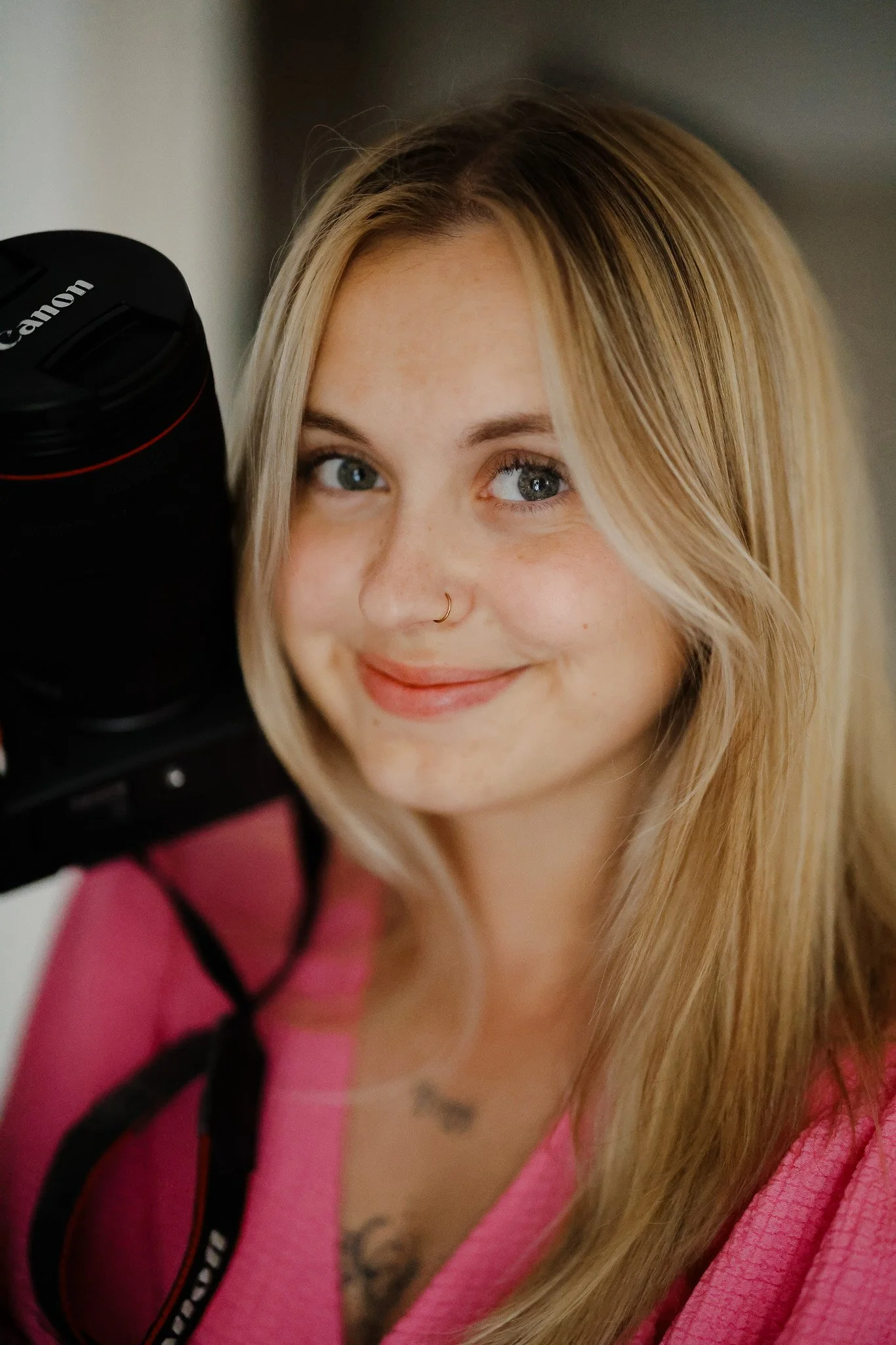 Une jeune femme aux cheveux blonds, portant un t-shirt rose, tenant un appareil photo Canon, souriante et regardant vers l'objectif.