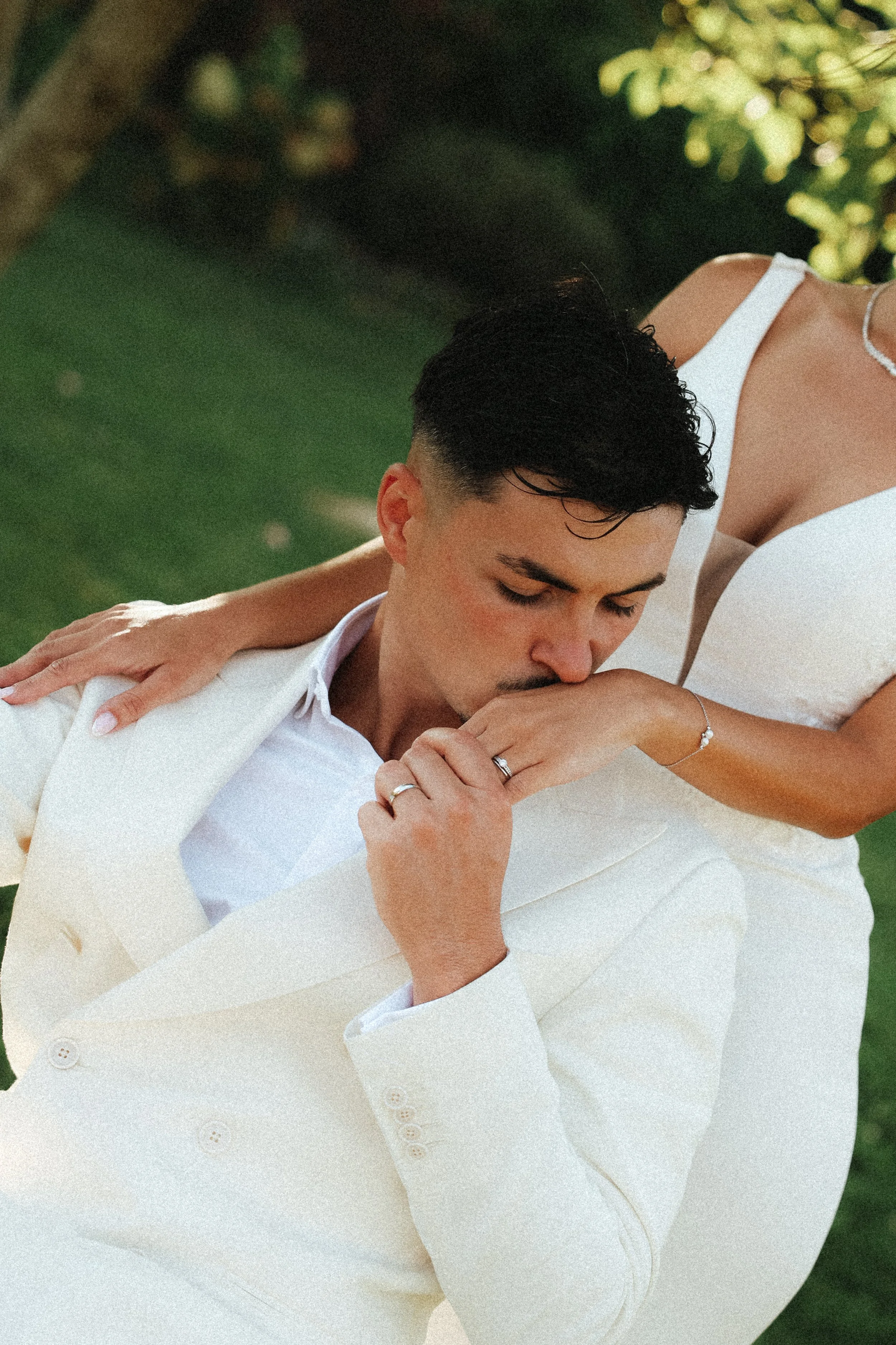 Un homme en costume blanc et une femme en robe blanche en extérieur, l'homme embrasse la main de la femme.