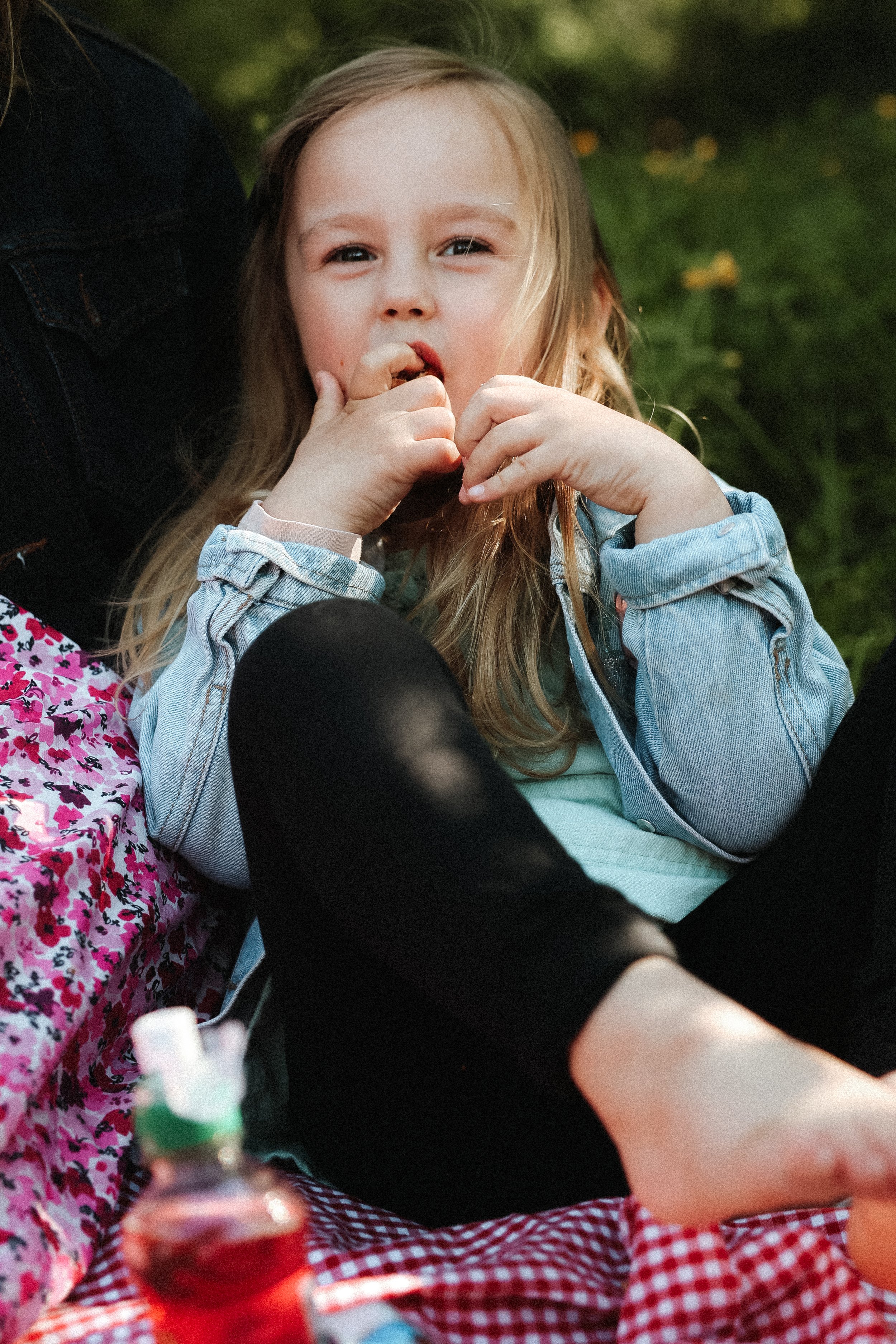 Une jeune fille blonde mange quelque chose et regarde vers l'appareil photo, assise à une table avec une nappe à carreaux rouges et blancs, en extérieur avec un fond de verdure.