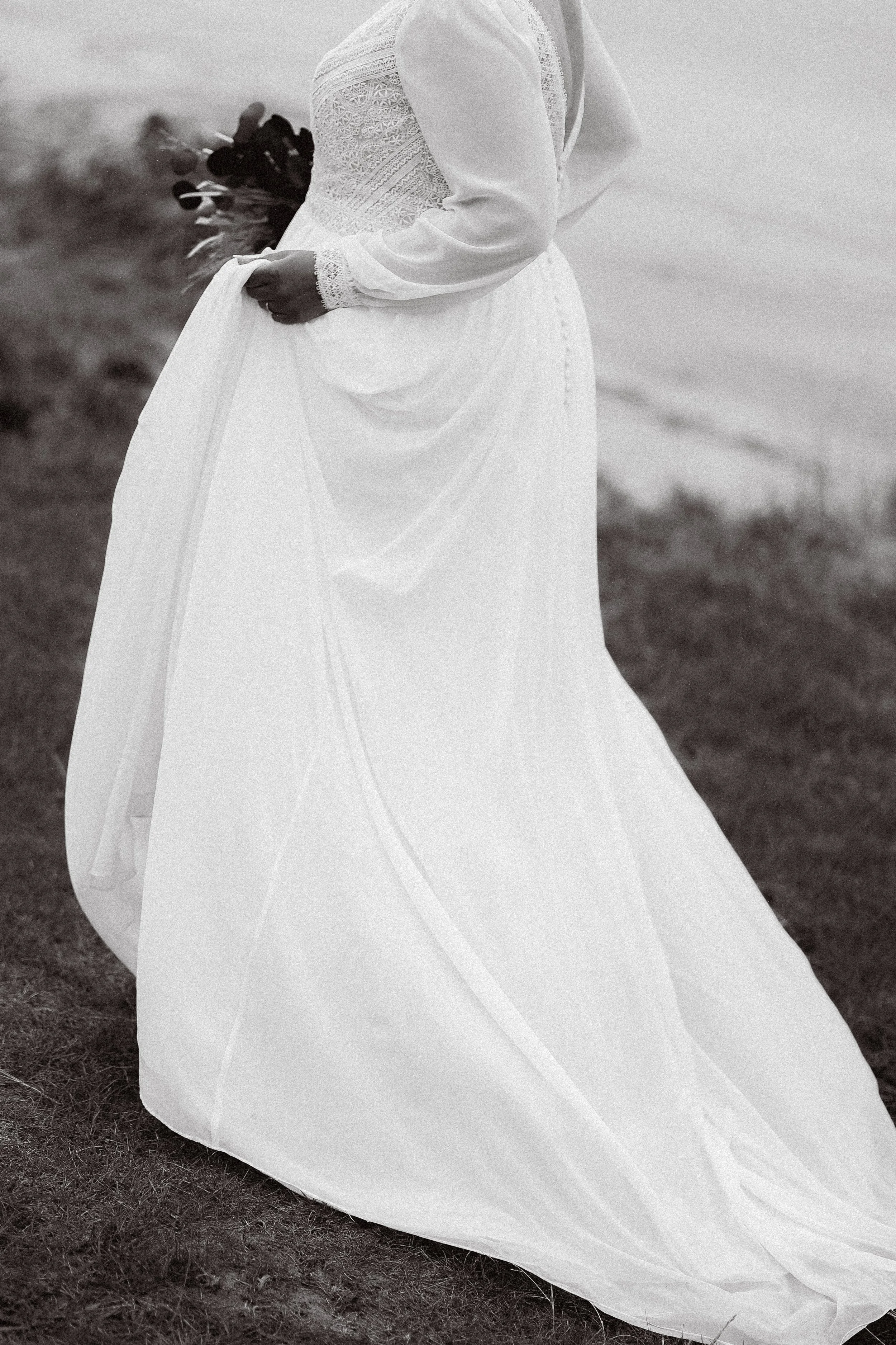 Une personne en robe de mariée blanche avec un bouquet, se tenant sur une plage ou un terrain herbeux.