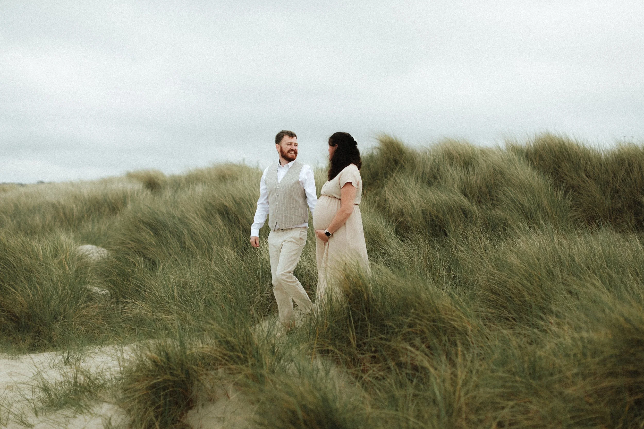 Un couple souriant lors d'une promenade dans un champ de hautes herbes, la femme est enceinte.